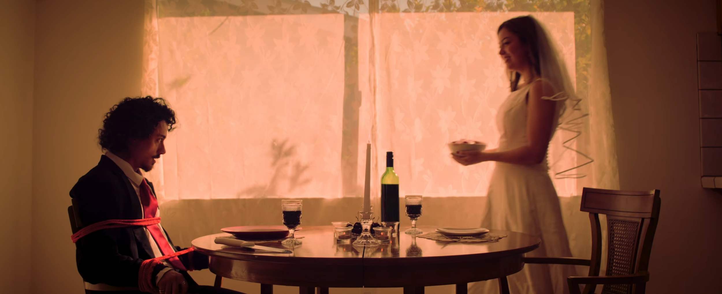 A man is tied to a chair at a dining table with red rope, while a woman stands nearby holding a bowl, smiling, in a warm-lit room with beige curtains.