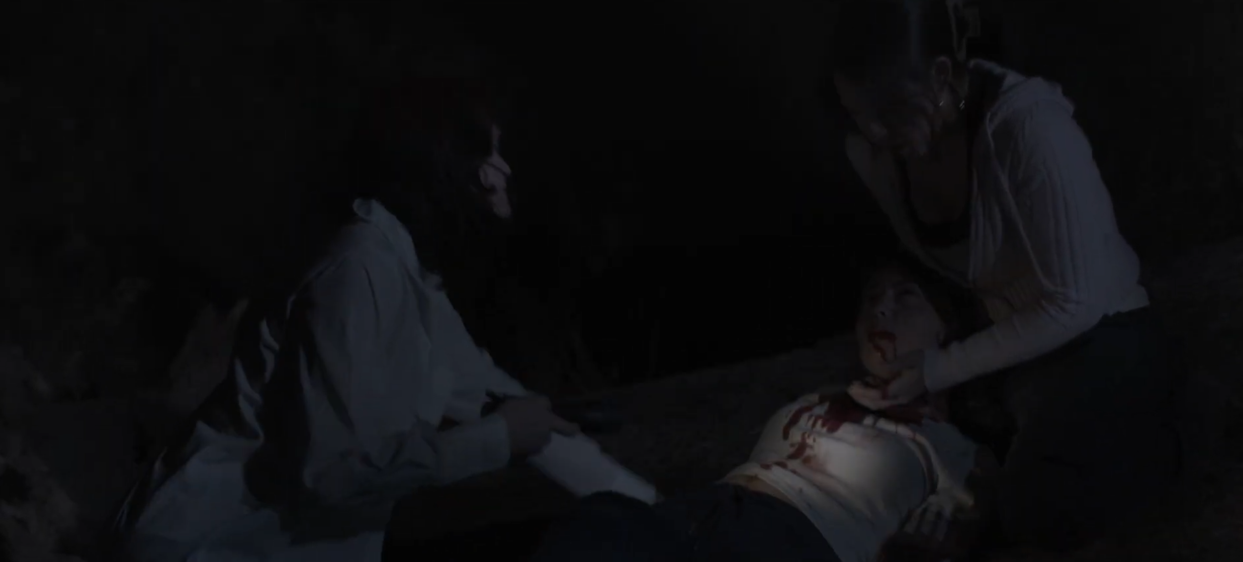 Three people in a dark environment, one appears to be injured and lying on the ground, while the others assist them.