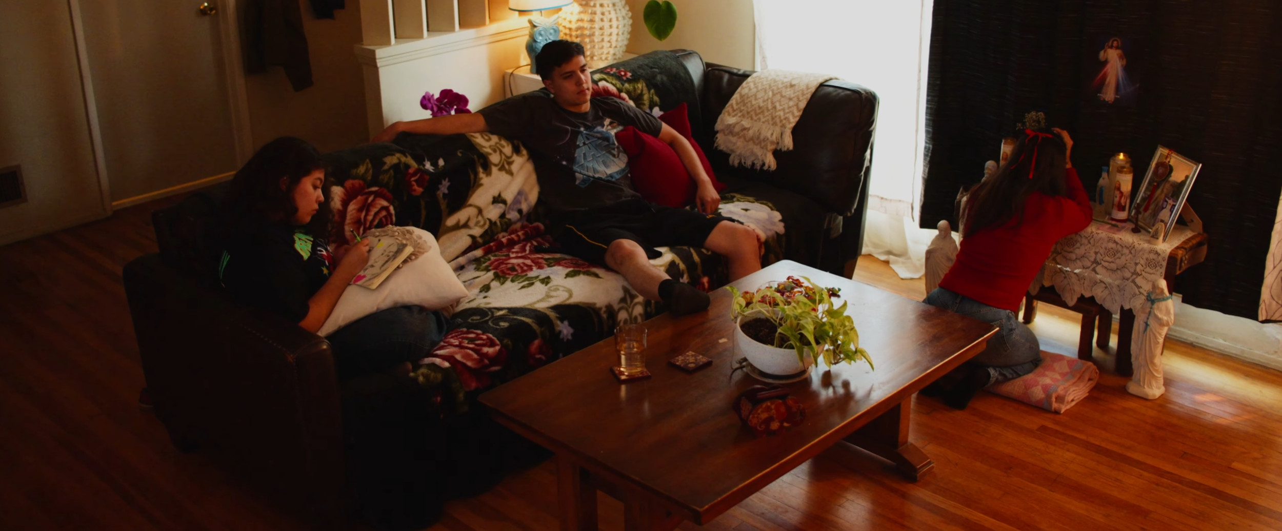 A living room scene with three children. A girl in a red top knelt at a small table near religious pictures and statues, a boy in a dark shirt and shorts sitting on a floral-patterned sofa, and another girl with dark hair reading a book on a pillow on the sofa. There is a wooden coffee table with a potted plant and some small items, and a window with light curtains in the background.