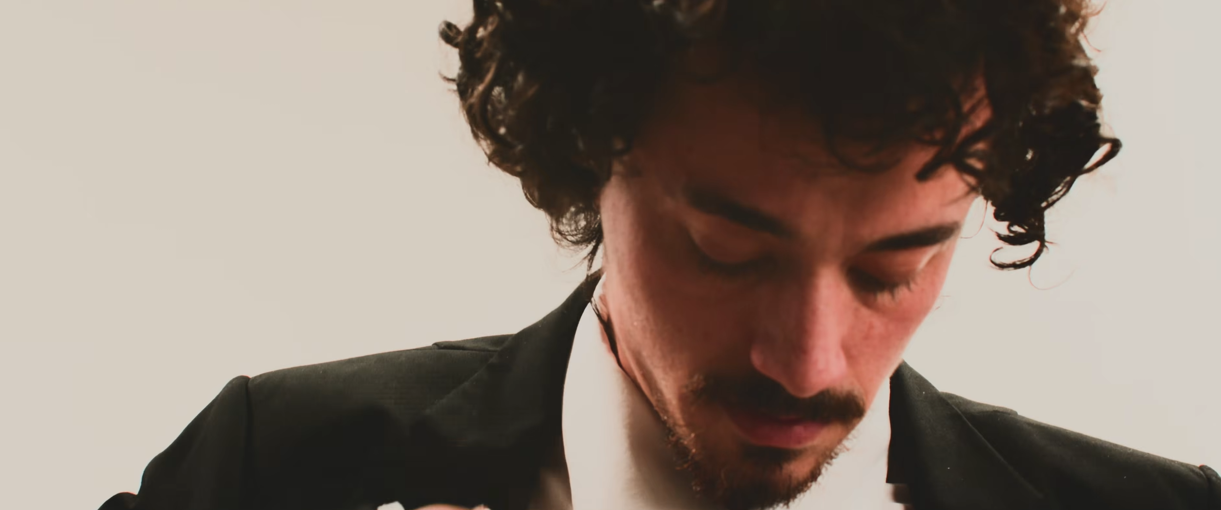Close-up of a man with curly hair and facial hair looking downward, wearing a black suit and white shirt against a plain light background.