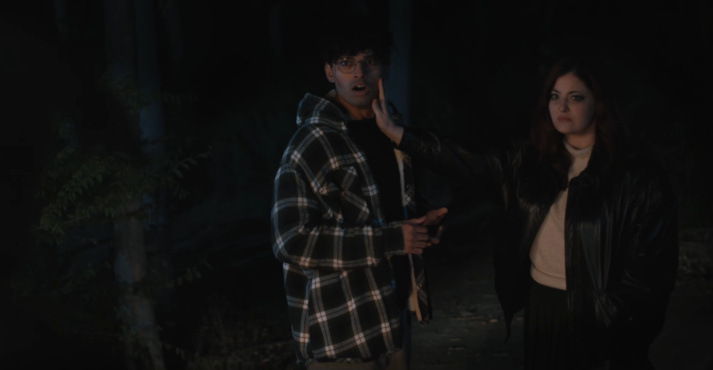 A man in glasses and a plaid jacket looks surprised while another woman with dark hair and a leather jacket touches his face in an outdoor setting at night.