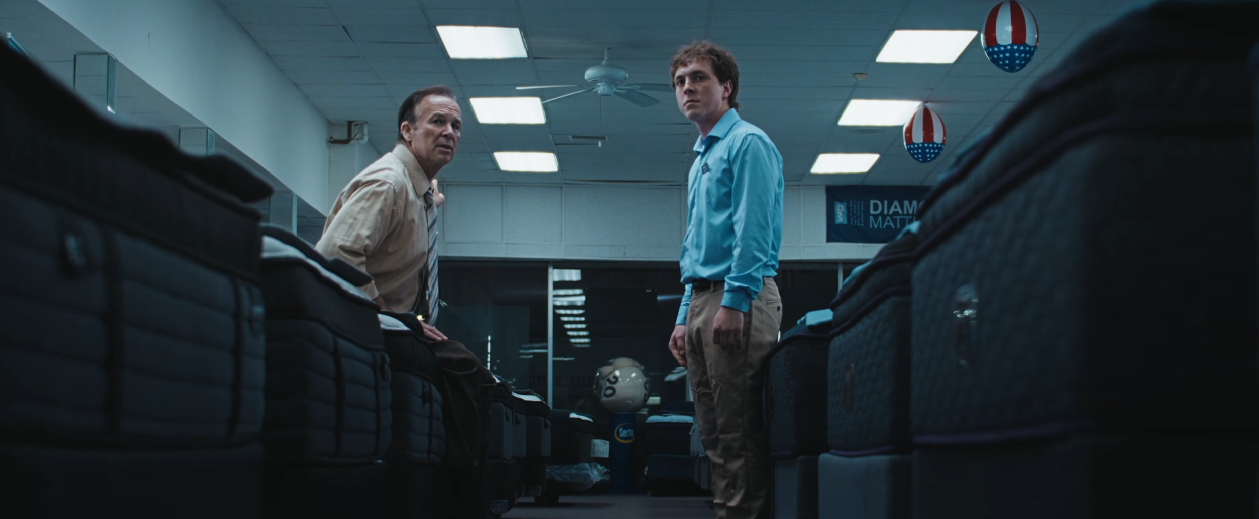 Two men standing in an airport store surrounded by bags and backpacks, with an American flag-themed balloon hanging from the ceiling.
