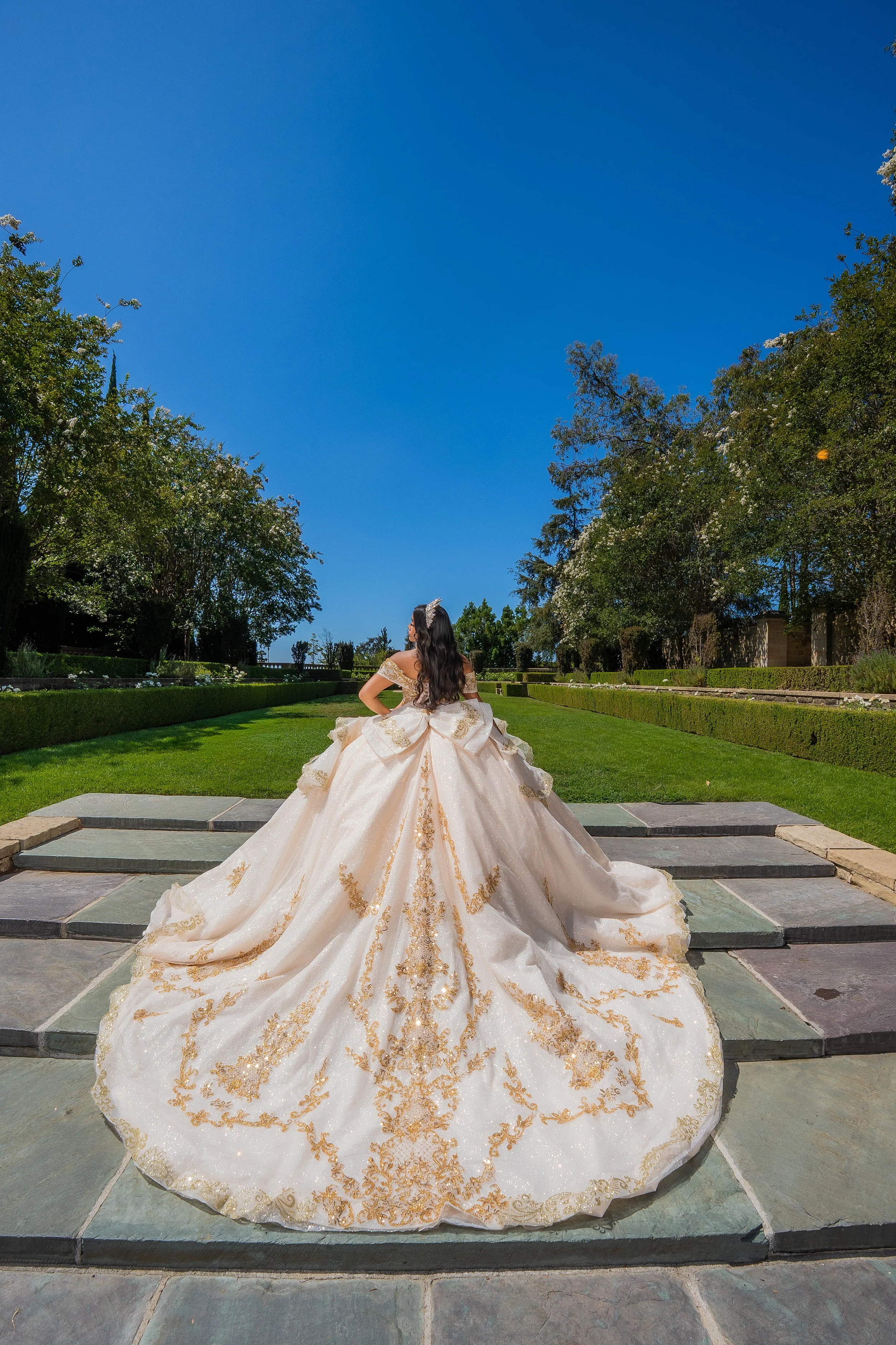 A woman in a white and gold ornate gown sitting on steps outside in a lush garden with trees and a clear blue sky.