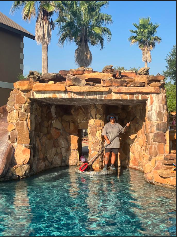 Person standing inside a stone-covered pool or water feature, holding a cleaning tool. Behind them are palm trees and a clear blue sky.