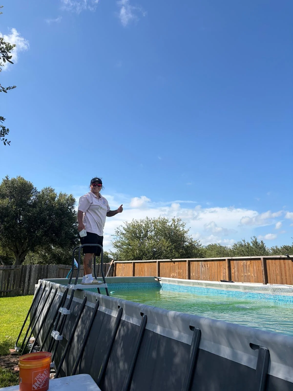 A person standing on a ladder in an above-ground swimming pool holding a cup, with a backyard and blue sky in the background.