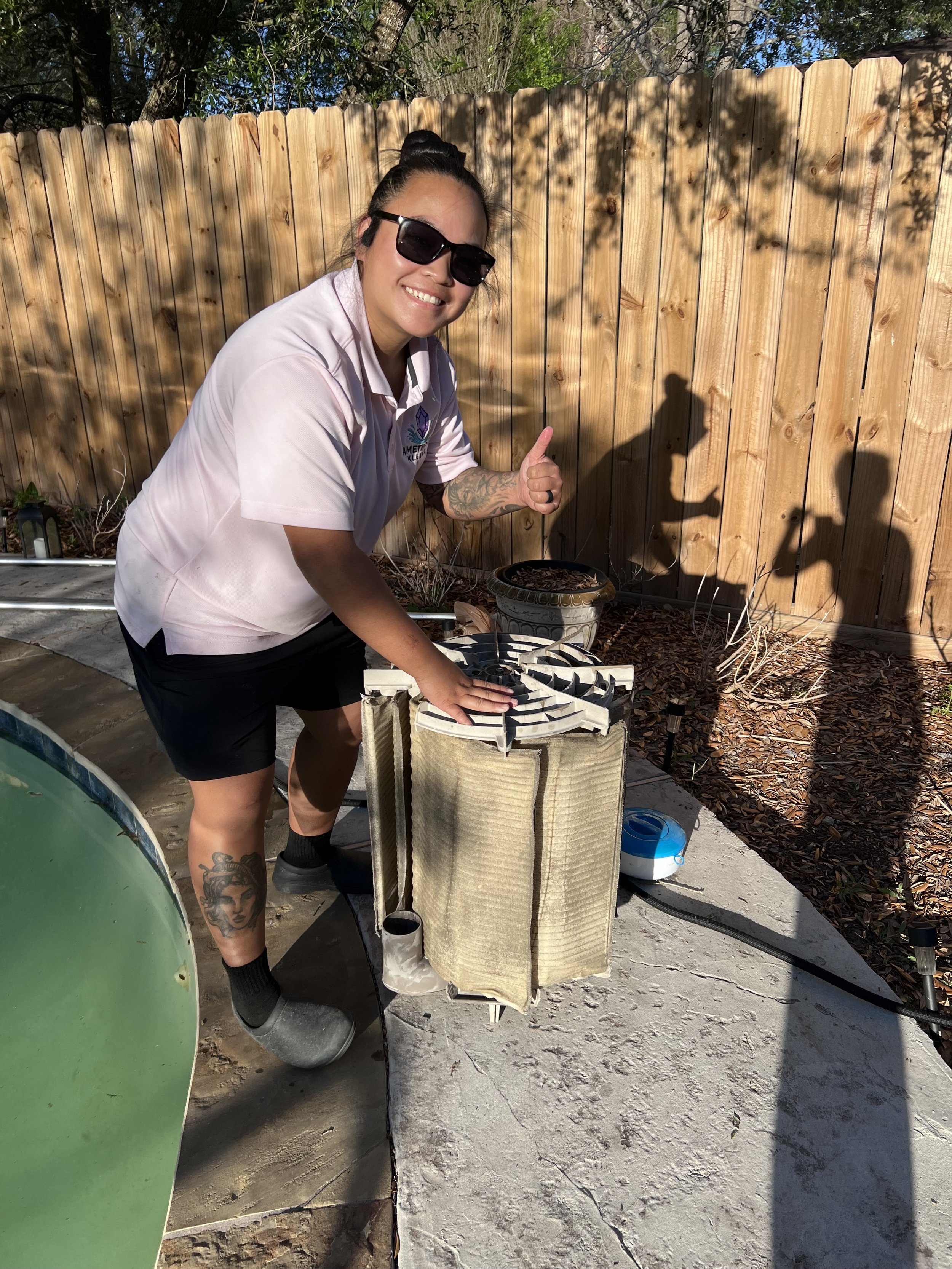 A woman with dark hair in a bun, wearing sunglasses, a white shirt, and black shorts, standing outdoors next to a makeshift water filter. She is giving a thumbs up and smiling. There is a wooden fence and trees in the background, with sunlight casting shadows.