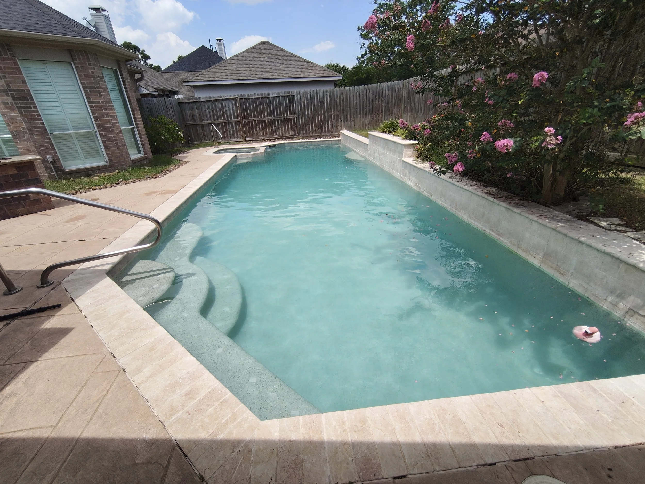 A backyard swimming pool surrounded by a patio, brick house, wooden fence, and flowering bushes, with a pink float toy in the pool.