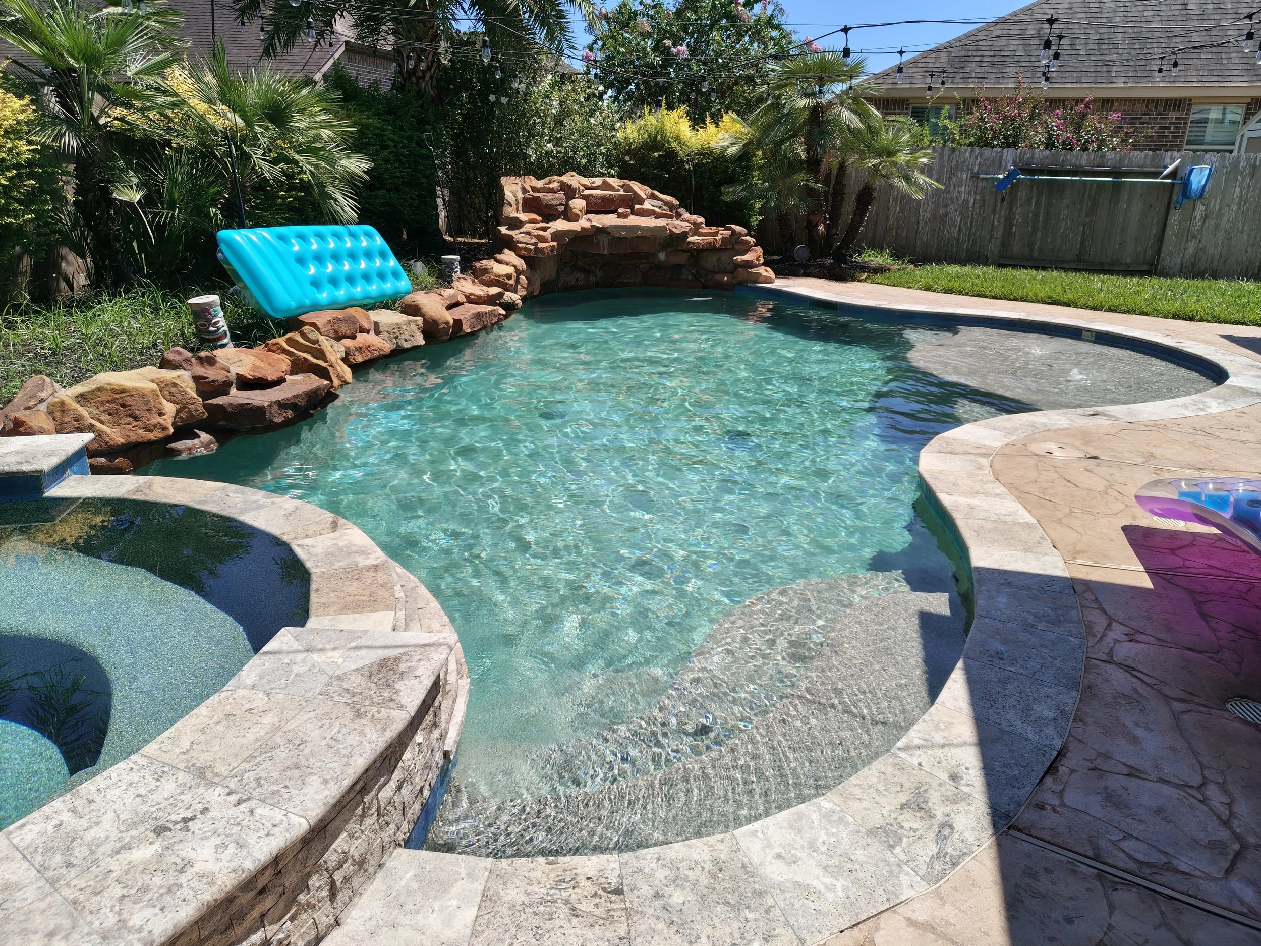 A backyard swimming pool area with a stone waterfall feature, surrounded by lush green plants and trees, with a wooden fence and a house in the background. Pool floatation devices are visible on the edge.