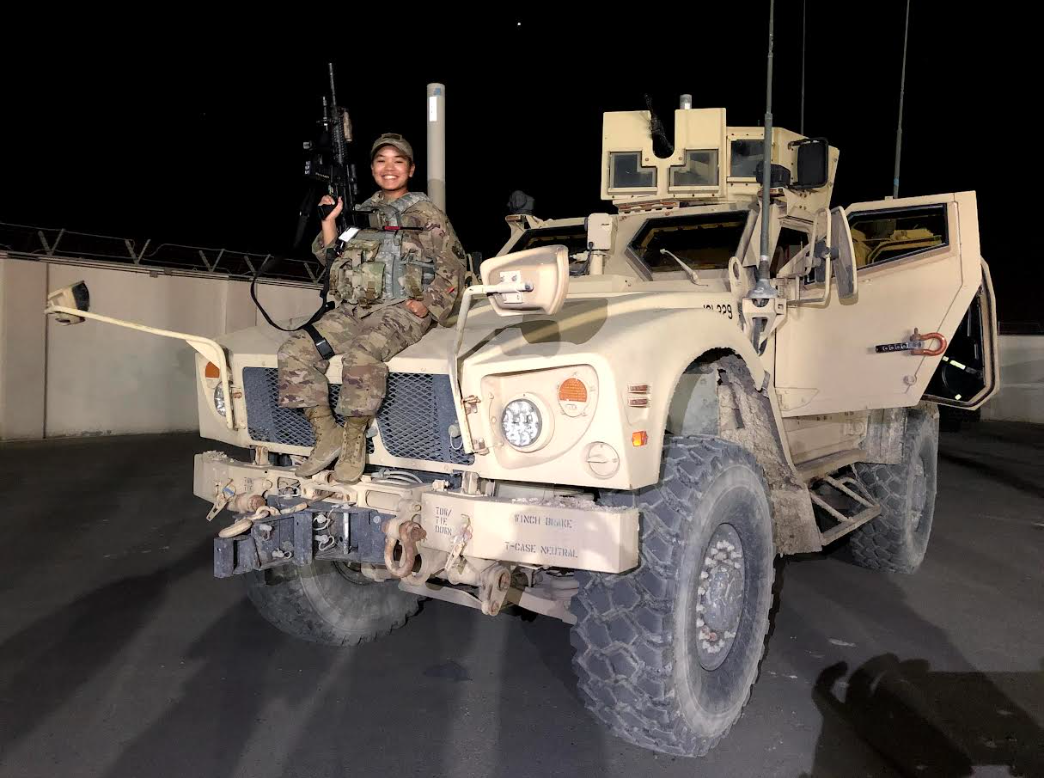 A smiling female soldier in military uniform sitting on top of a large beige military vehicle at night, holding a rifle, with a dark sky and a concrete barrier in the background.