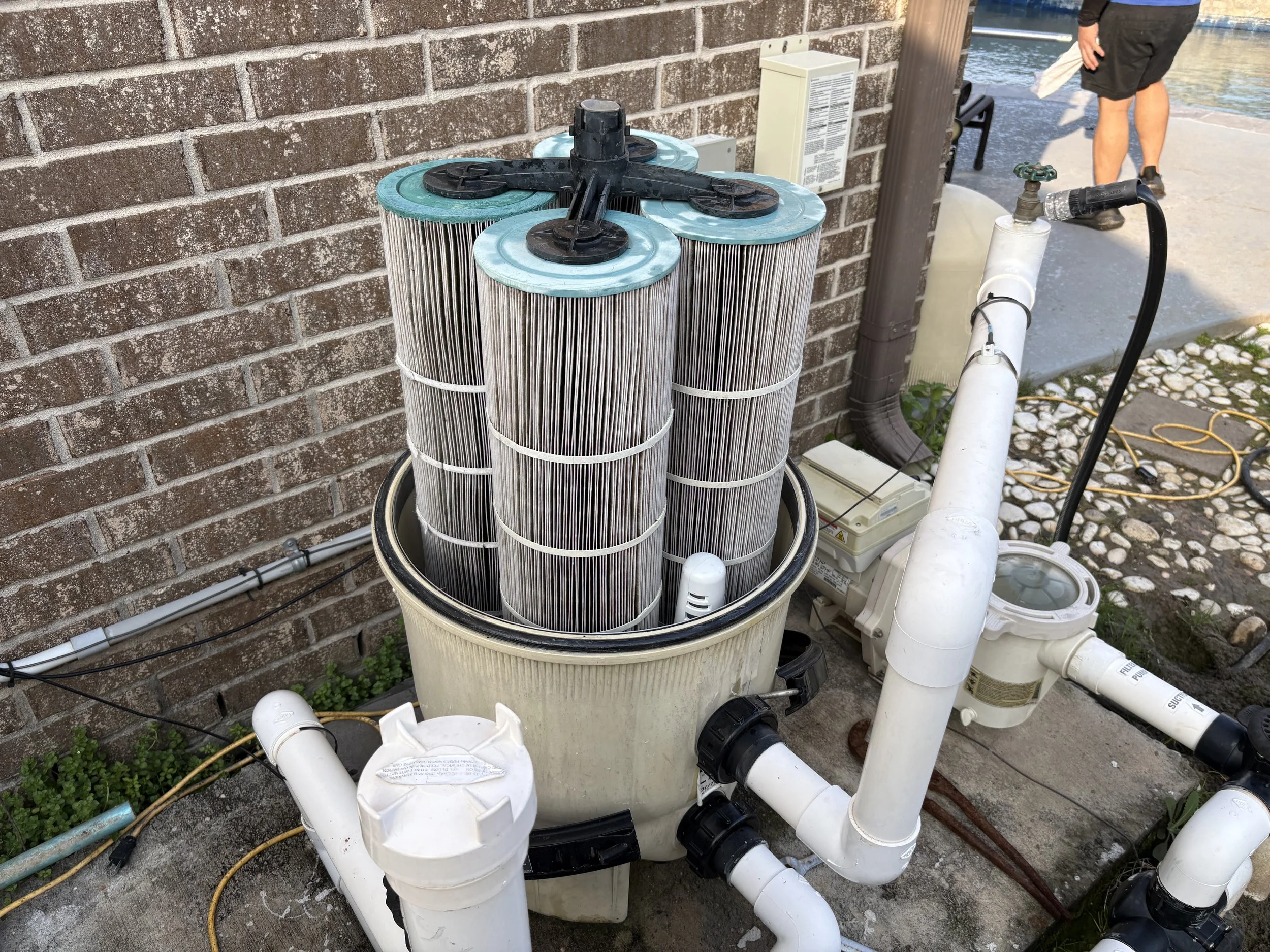 Pool filter system with four large cylindrical filters mounted on a container, surrounded by white PVC pipes connected to pool equipment, next to a brick wall and near a water body.