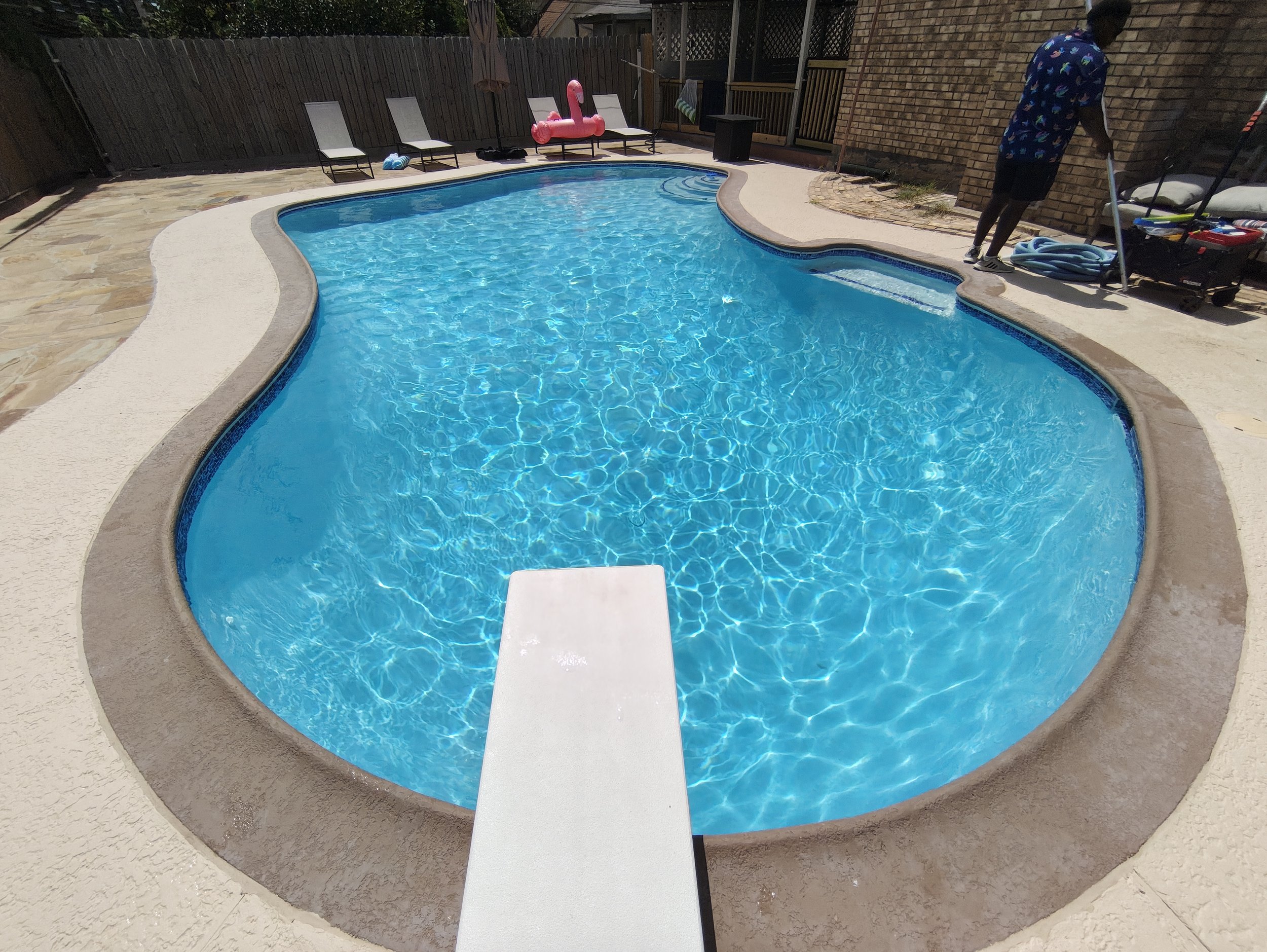 In-ground backyard swimming pool with blue water, white diving board, pink flamingo pool float, poolside chairs, man in tropical shirt and black shorts near wall, hose and pool cleaning tools on side.