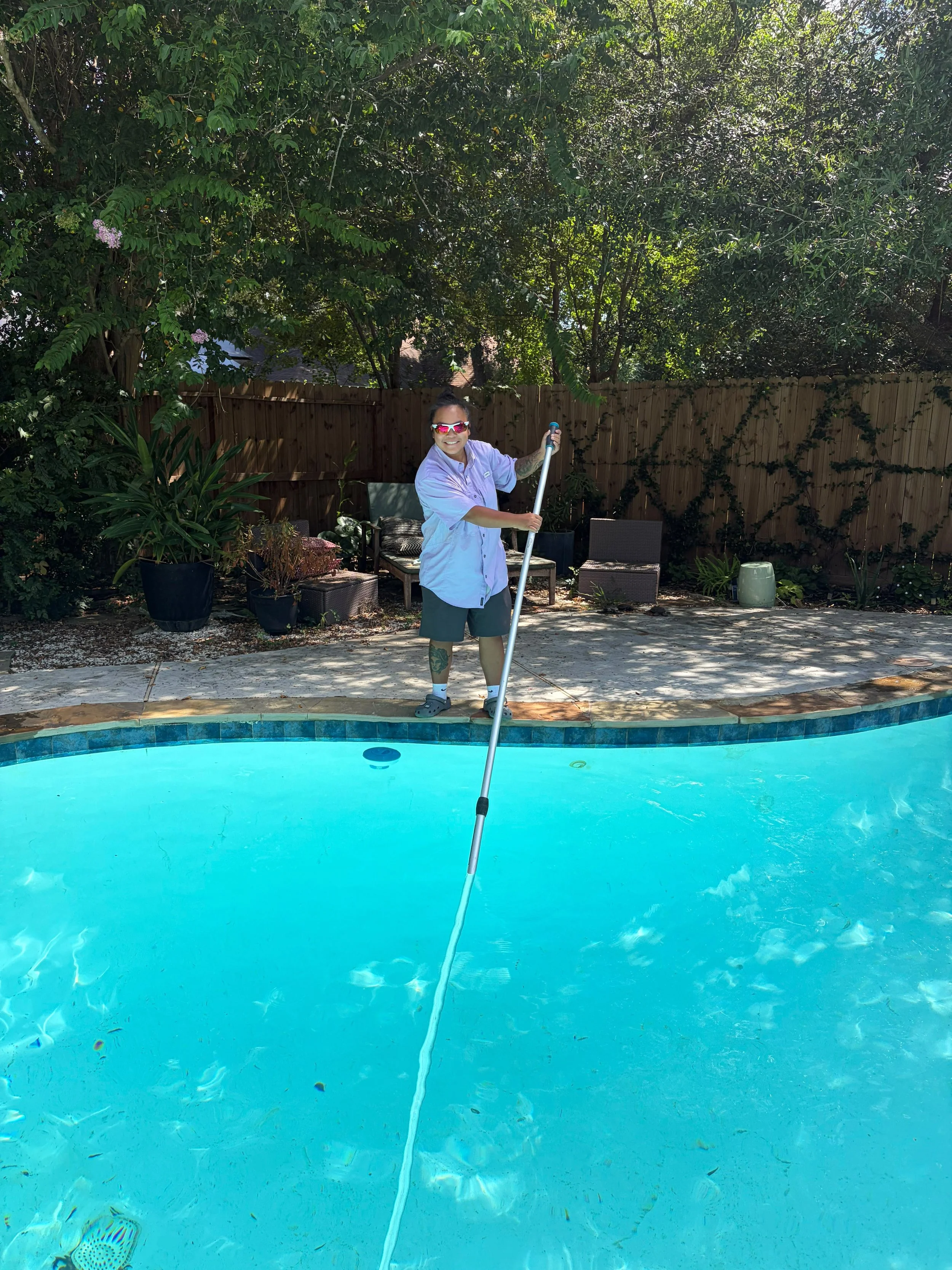 A person standing next to a swimming pool, holding a pool skimmer, smiling, wearing sunglasses, a light blue short-sleeved shirt, dark shorts, and Crocs, with a wooden fence and leafy trees in the background.