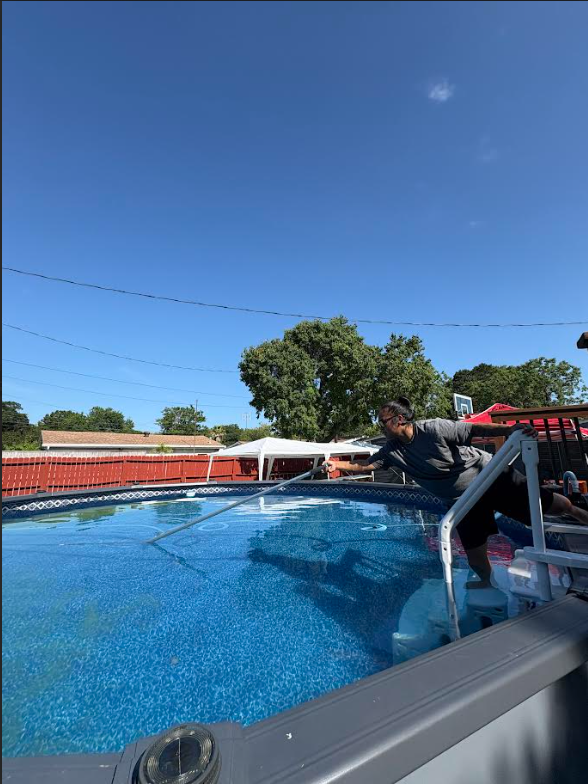 A person cleaning or inspecting an above-ground swimming pool with a long pole, pool is filled with water, backyard with trees, fence, and a basketball hoop in the background, clear blue sky.
