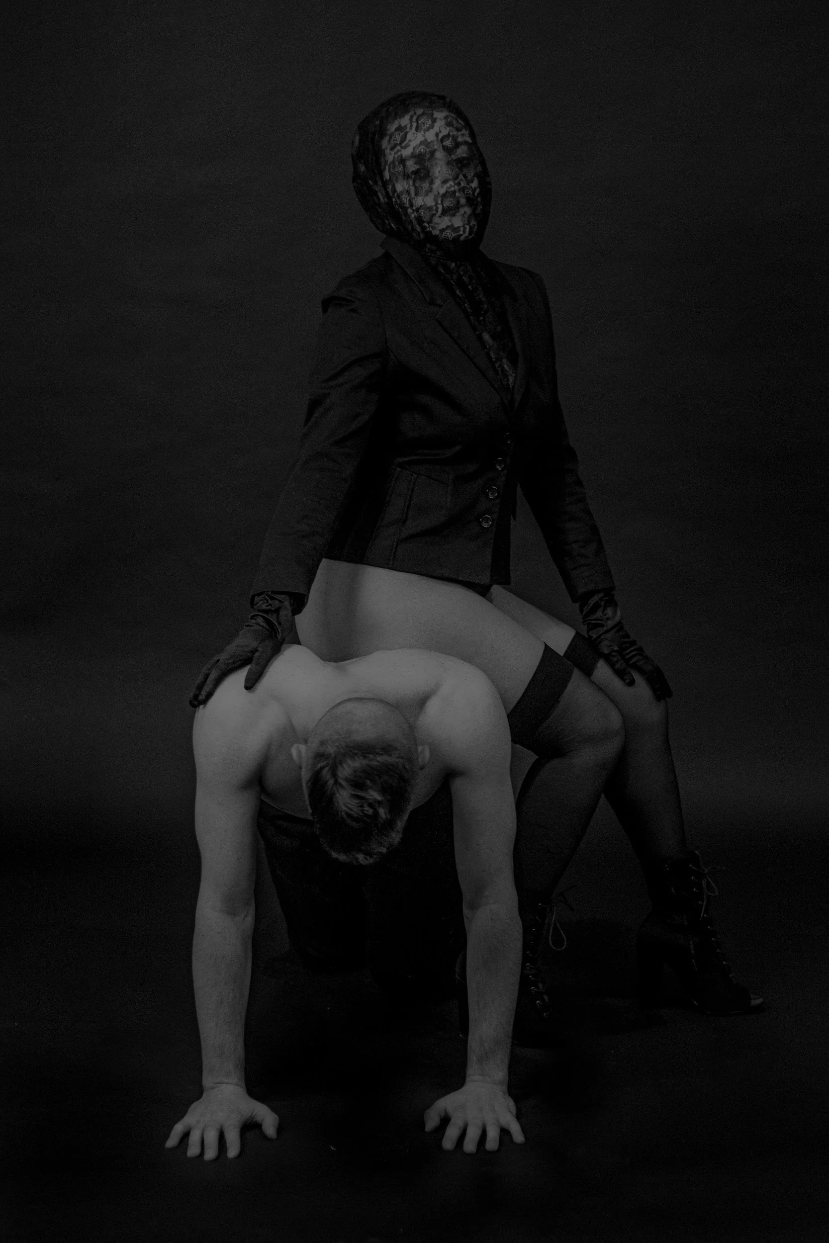 A person with a lace mask and black outfit sitting on a woman bent forward on all fours, both posed against a dark background.