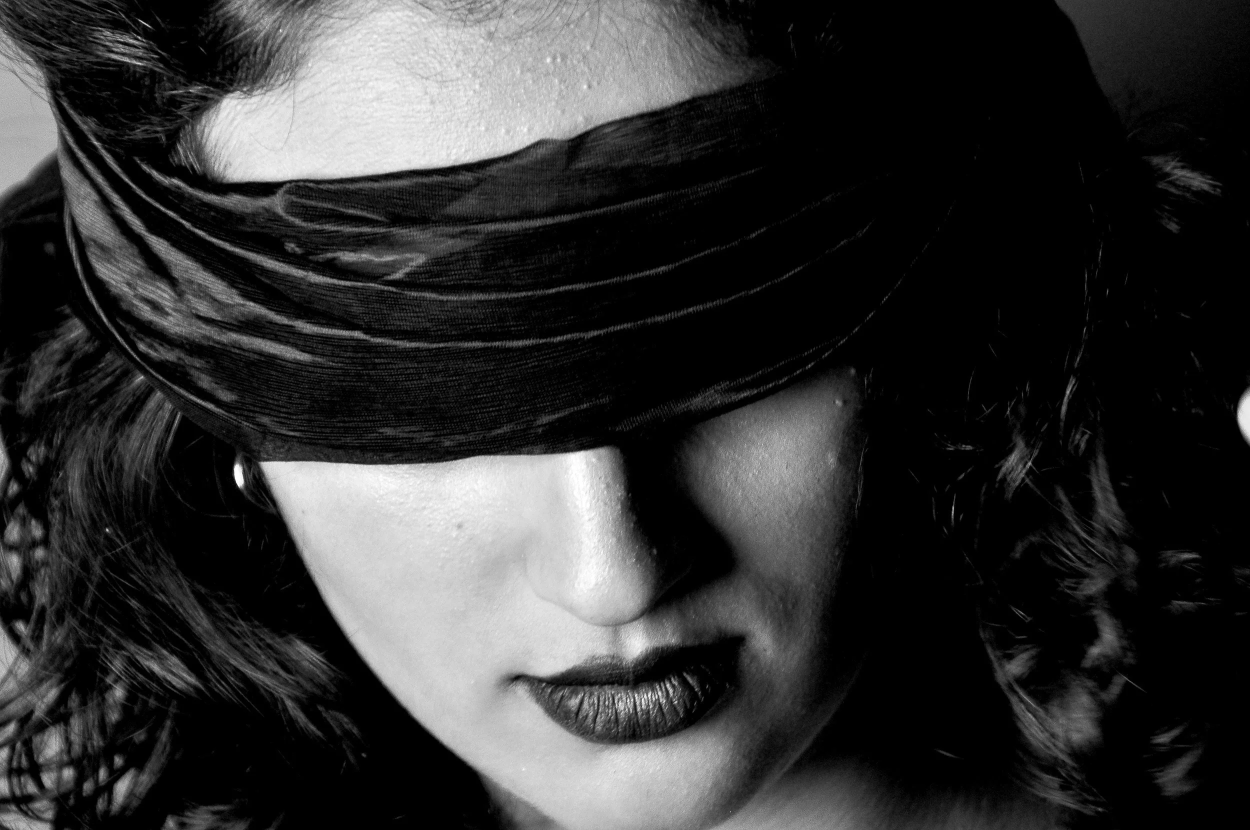 Close-up of a woman's face with makeup, her eyes covered by a textured fabric, showing her lips and part of her curly hair.