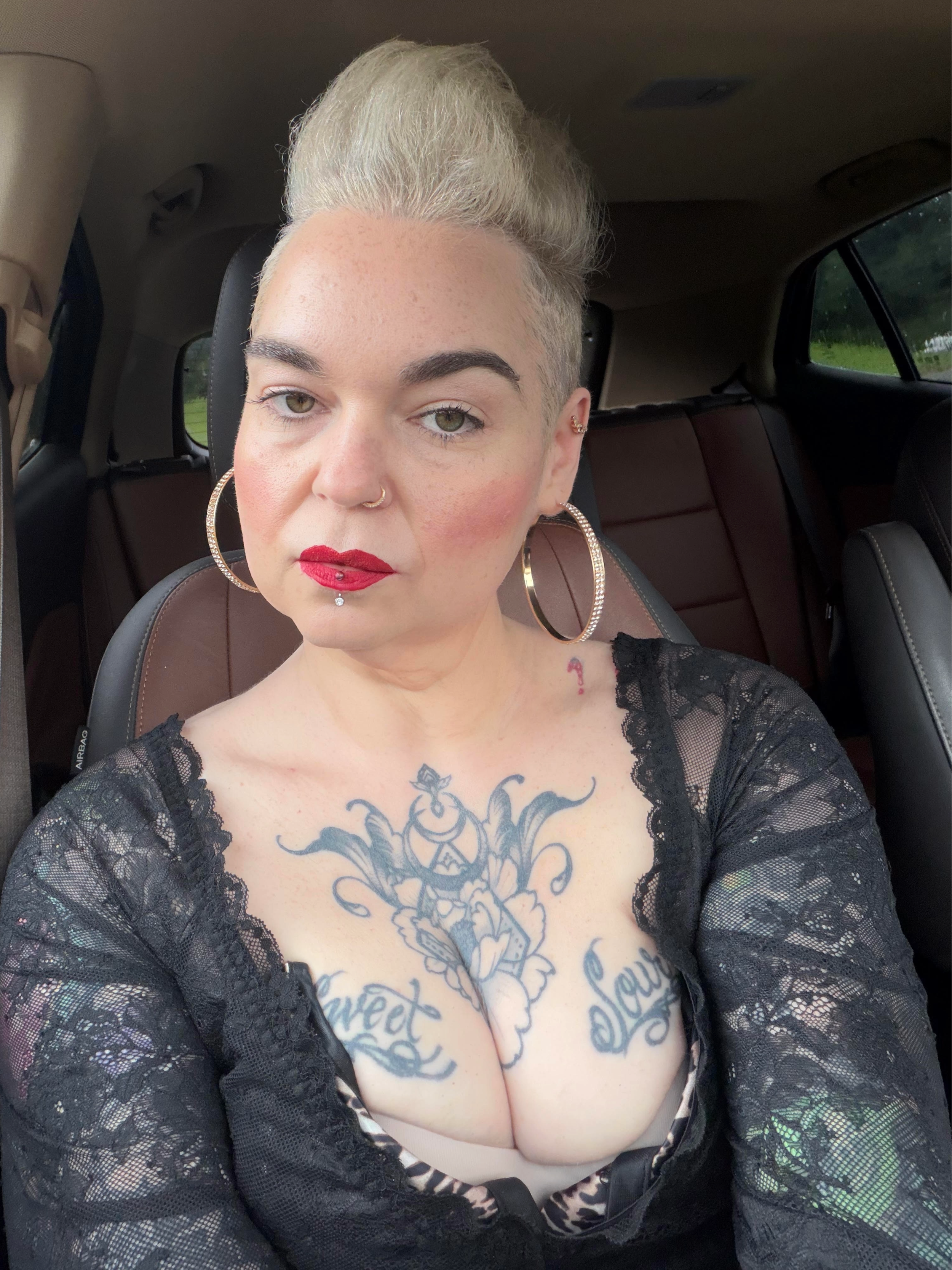 A woman with platinum blonde hair styled in a high pompadour, wearing makeup with bold red lipstick, large hoop earrings, a nose ring, and visible tattoos on her chest, sitting in a vehicle.