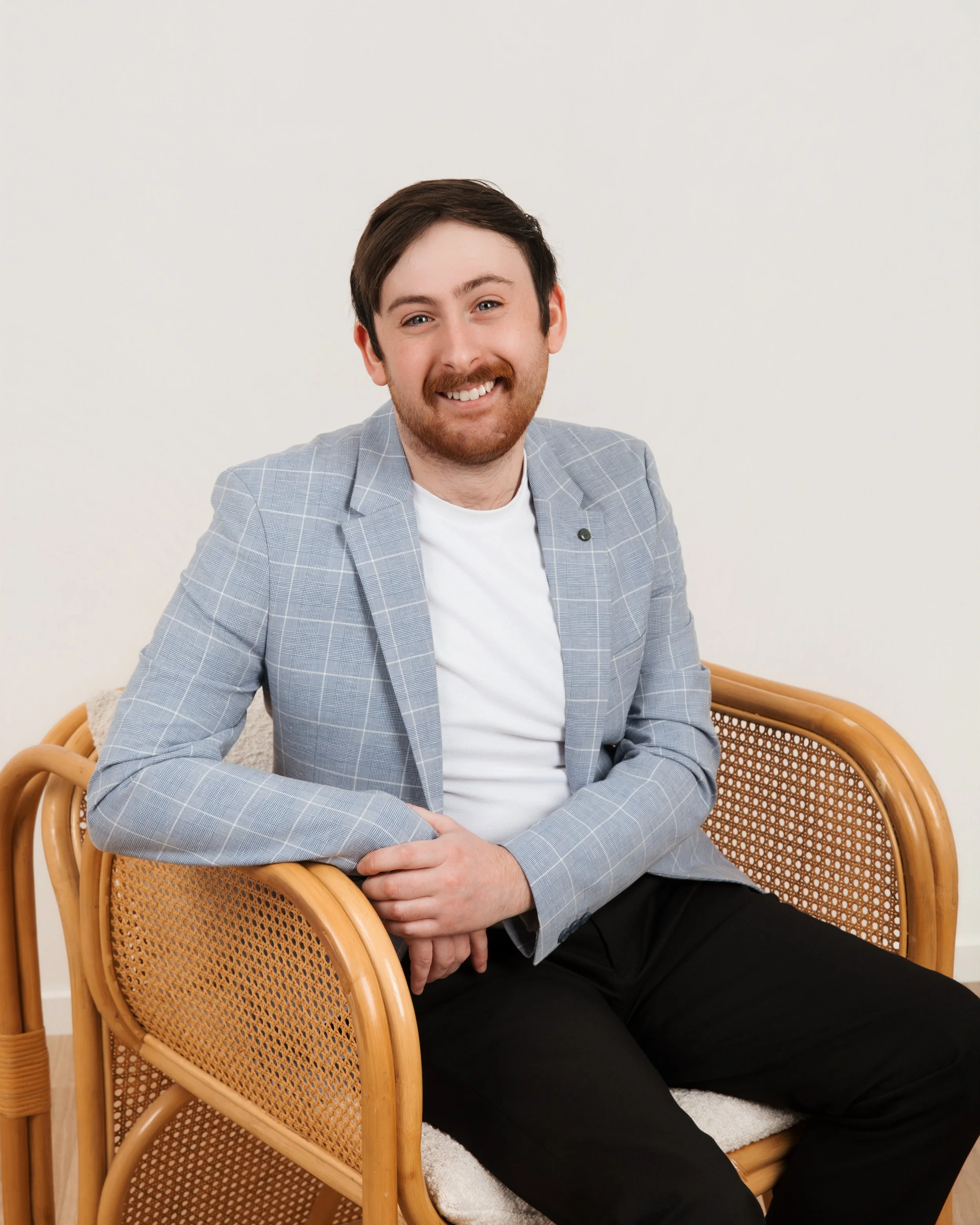 A young man with dark hair and a beard is sitting on a rattan chair with a white cushion, smiling at the camera. He is wearing a light gray checkered blazer over a white t-shirt and black pants. The background is plain white.