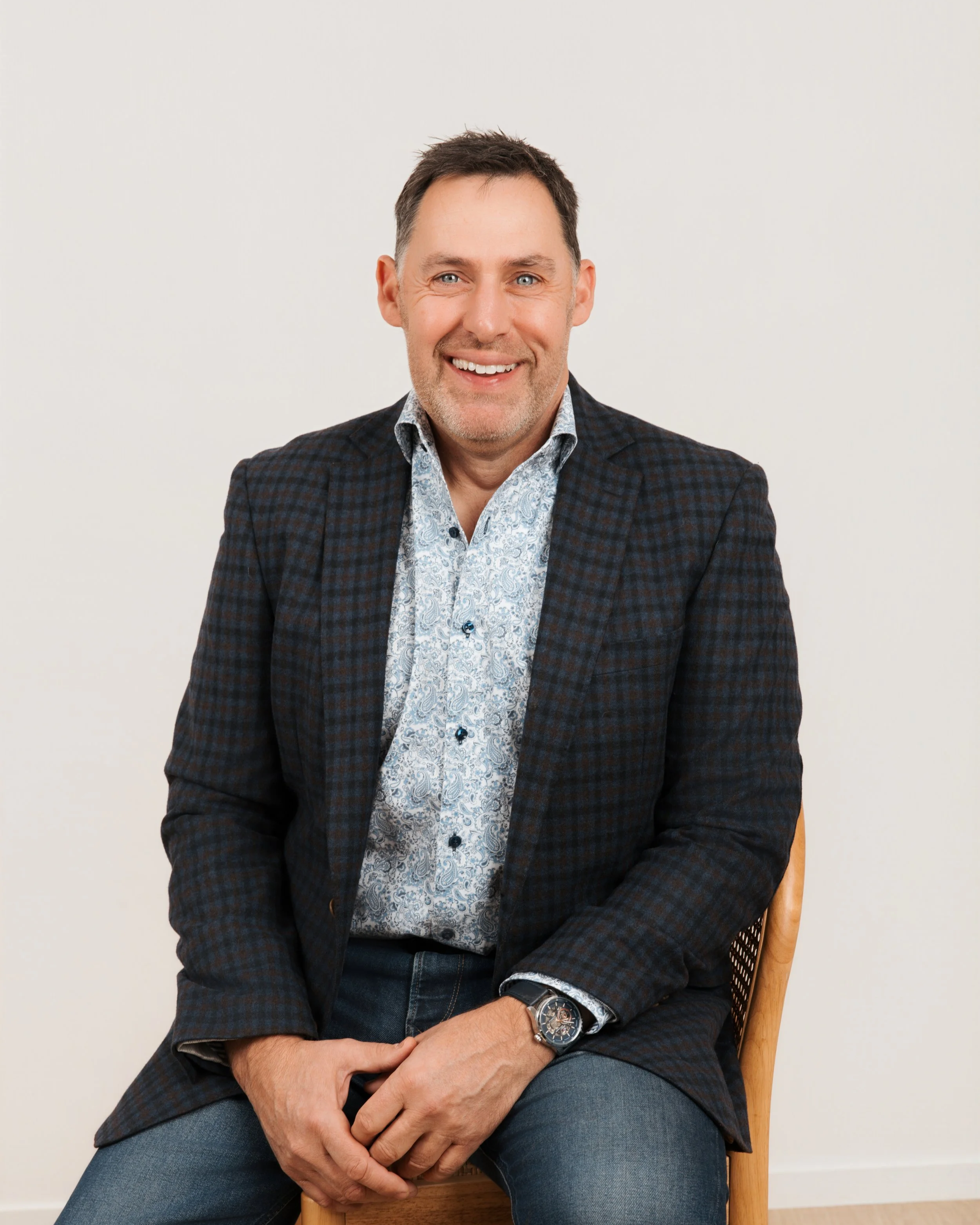 A man sitting on a wooden chair against a plain white background, wearing a patterned dress shirt and a dark blazer, smiling and looking at the camera.