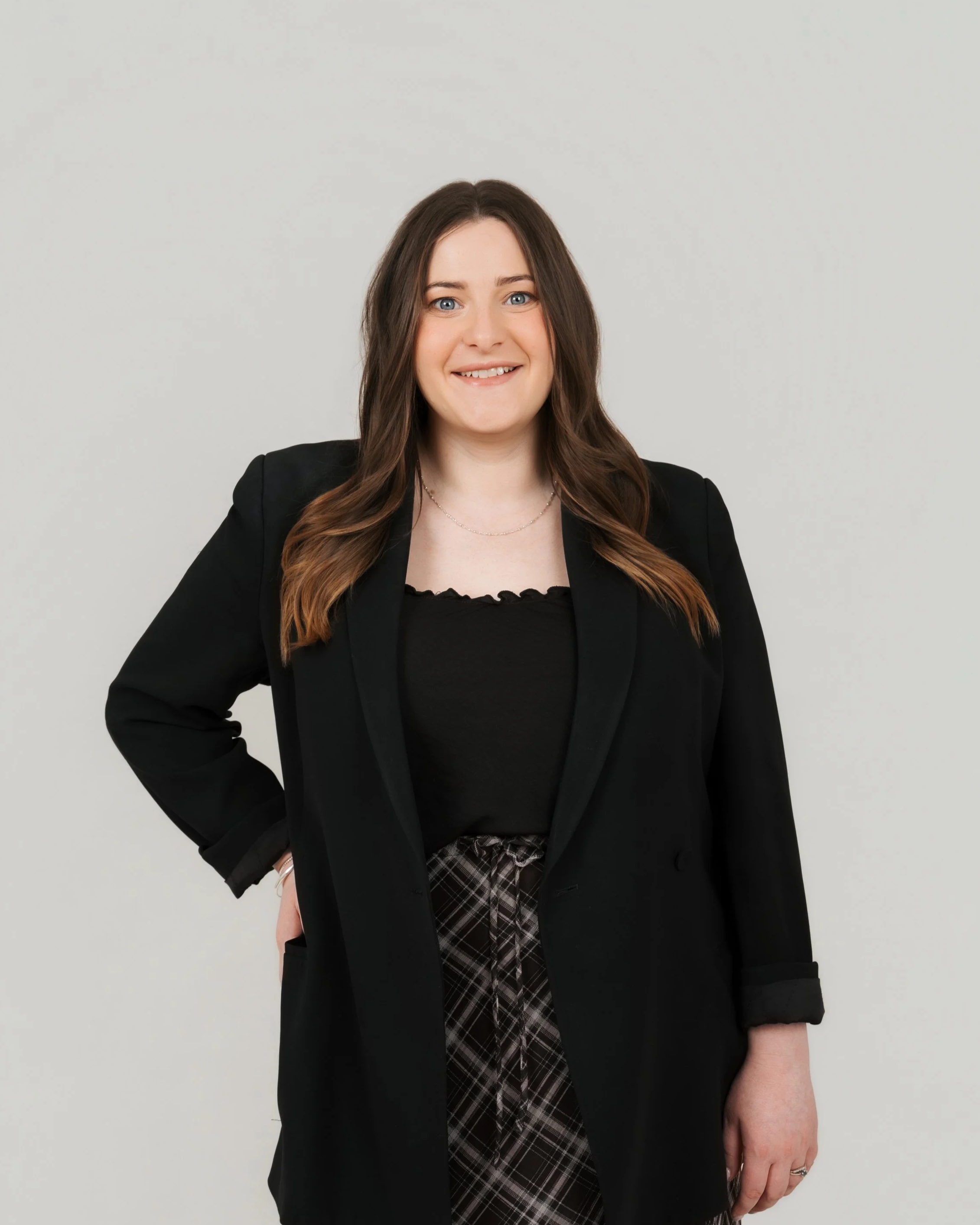 A woman with long brown hair, blue eyes, and a smile, wearing a black blazer, black top, and plaid pants, standing against a plain light gray background.