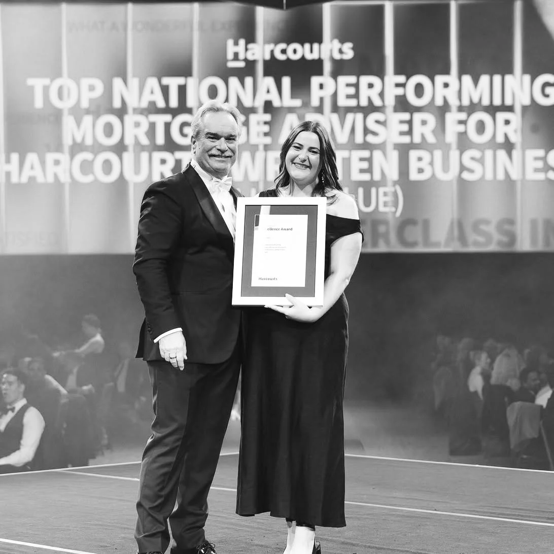 Two people in formal attire standing on stage, smiling, with a large award certificate between them, at an event celebrating Harcourts' achievements.