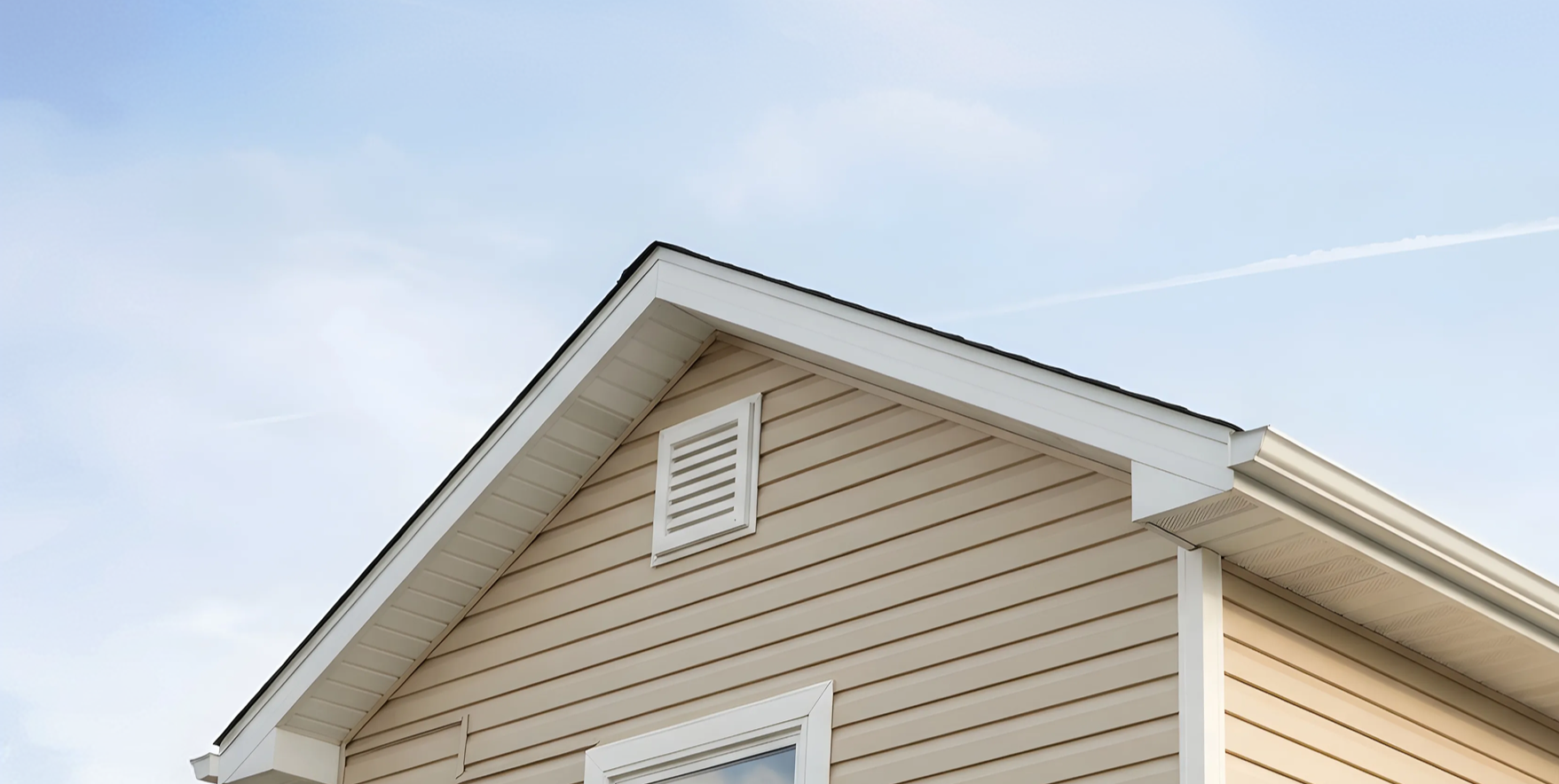 Close-up of a house's gable roof with beige siding, white trim, and a small attic vent, under a clear blue sky.