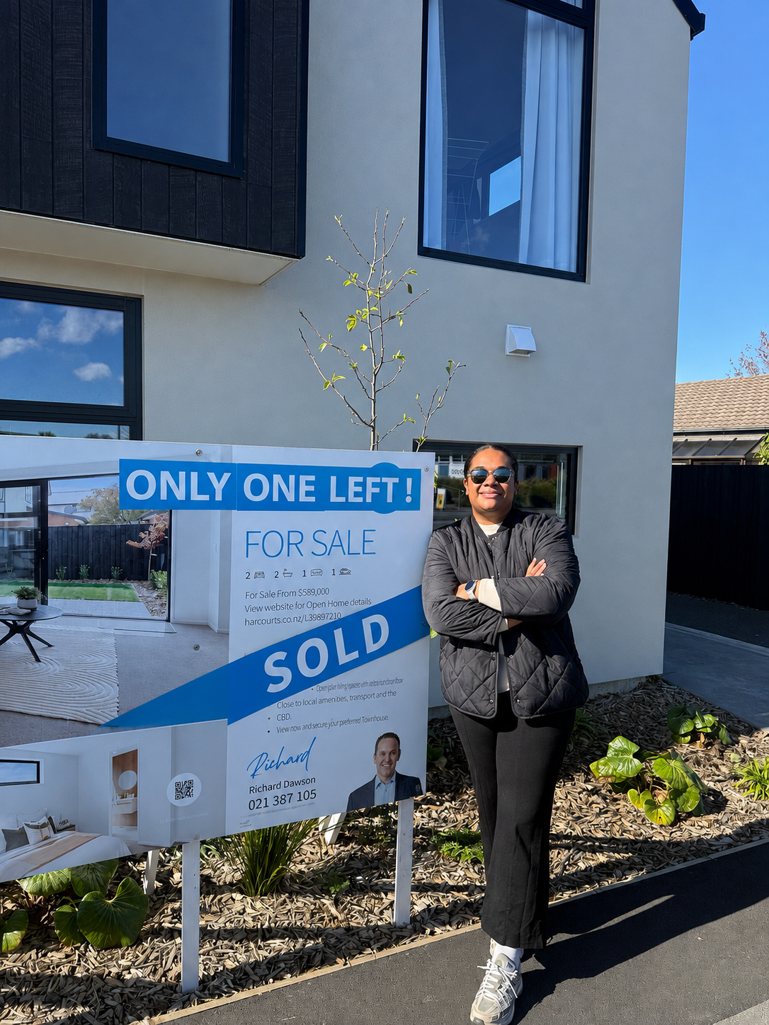 Woman in sunglasses standing beside a real estate sign that says 'Only One Left! Sold' in front of a modern house with large windows and a small tree.