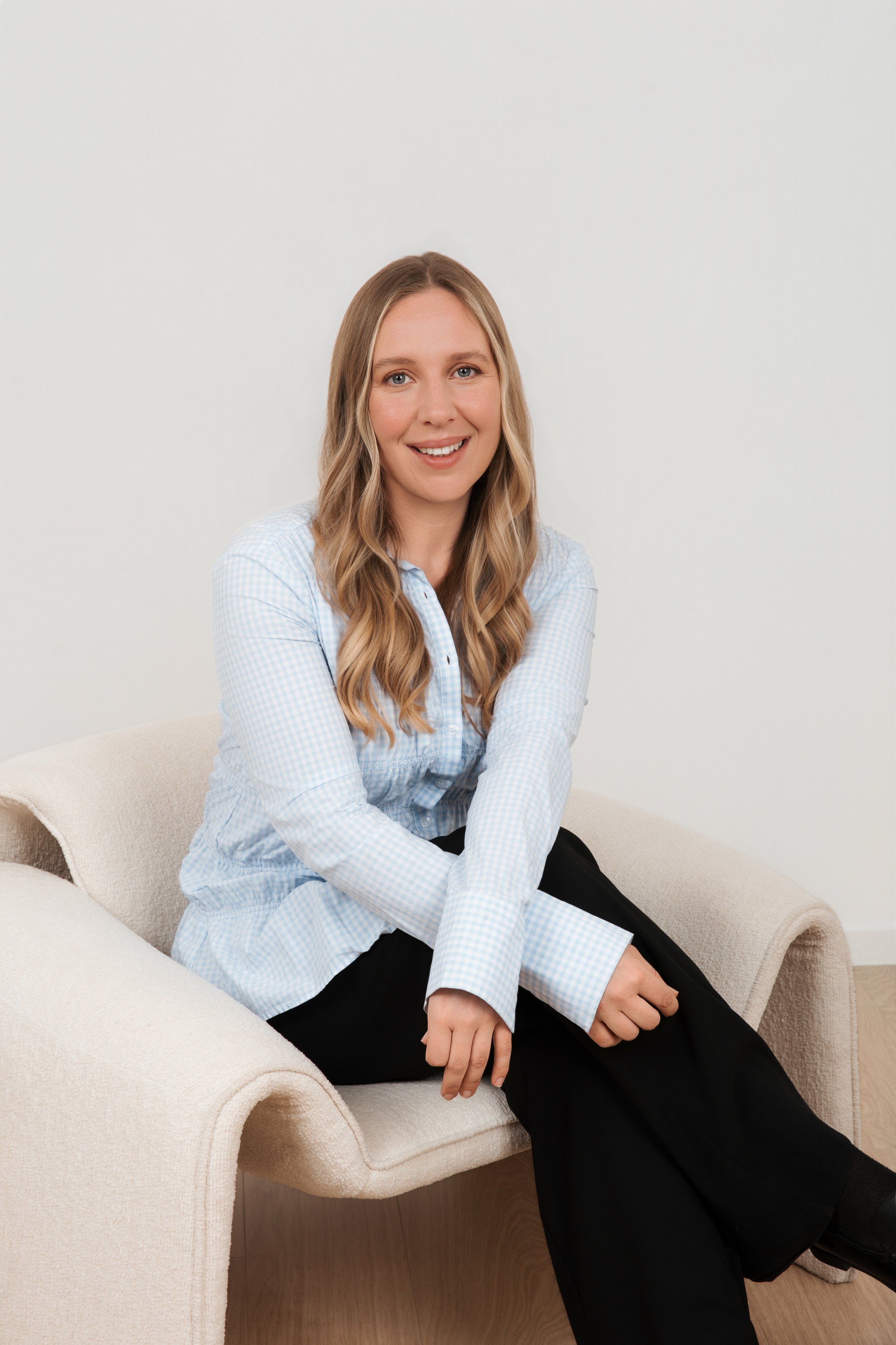 A woman with long blonde hair, wearing a light blue checked shirt and black pants, sitting on a beige couch, smiling at the camera against a plain white background.