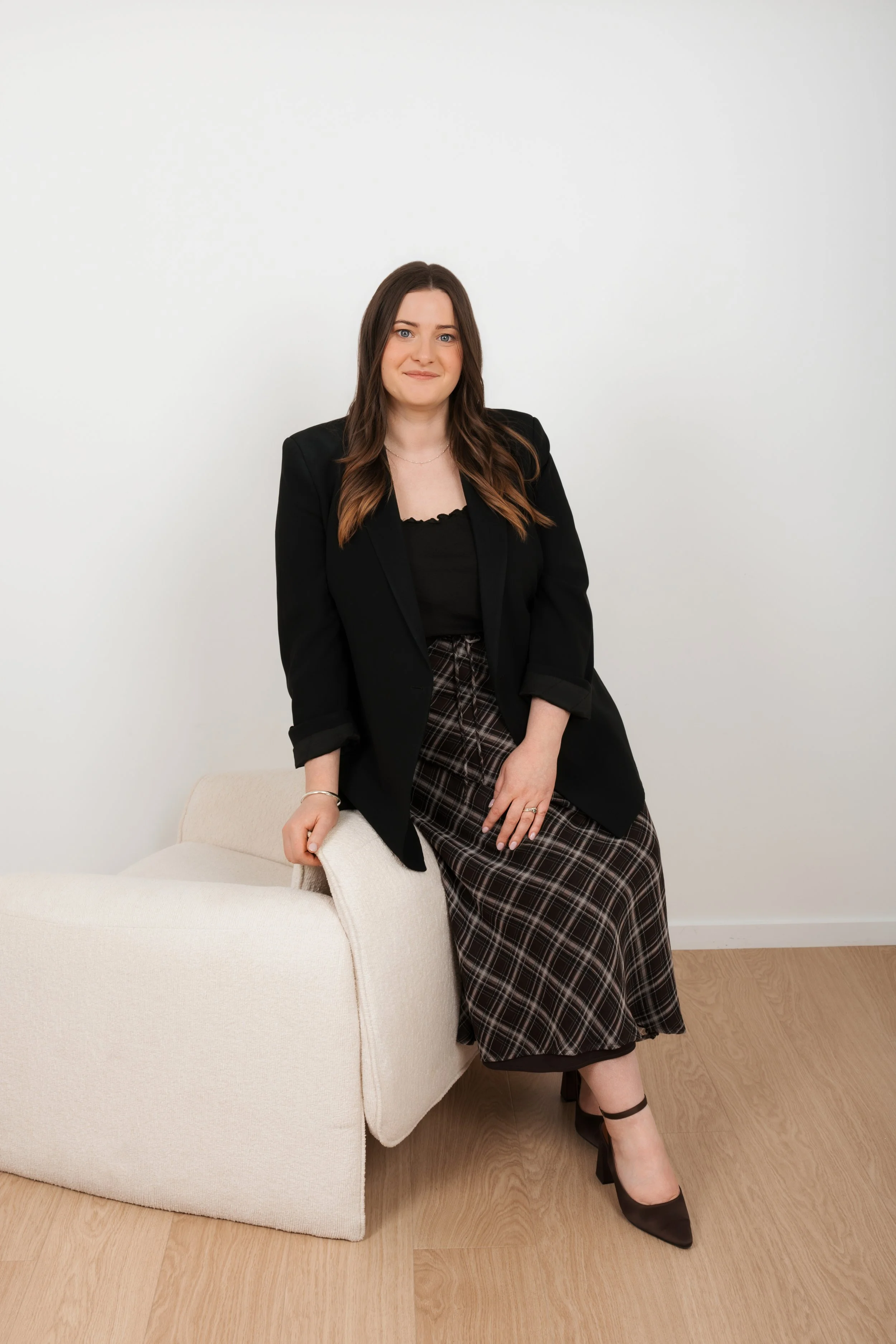 A woman with long brown hair, wearing a black blazer, black top, and plaid skirt, sitting on a white armchair in a room with wooden flooring and plain white wall background.