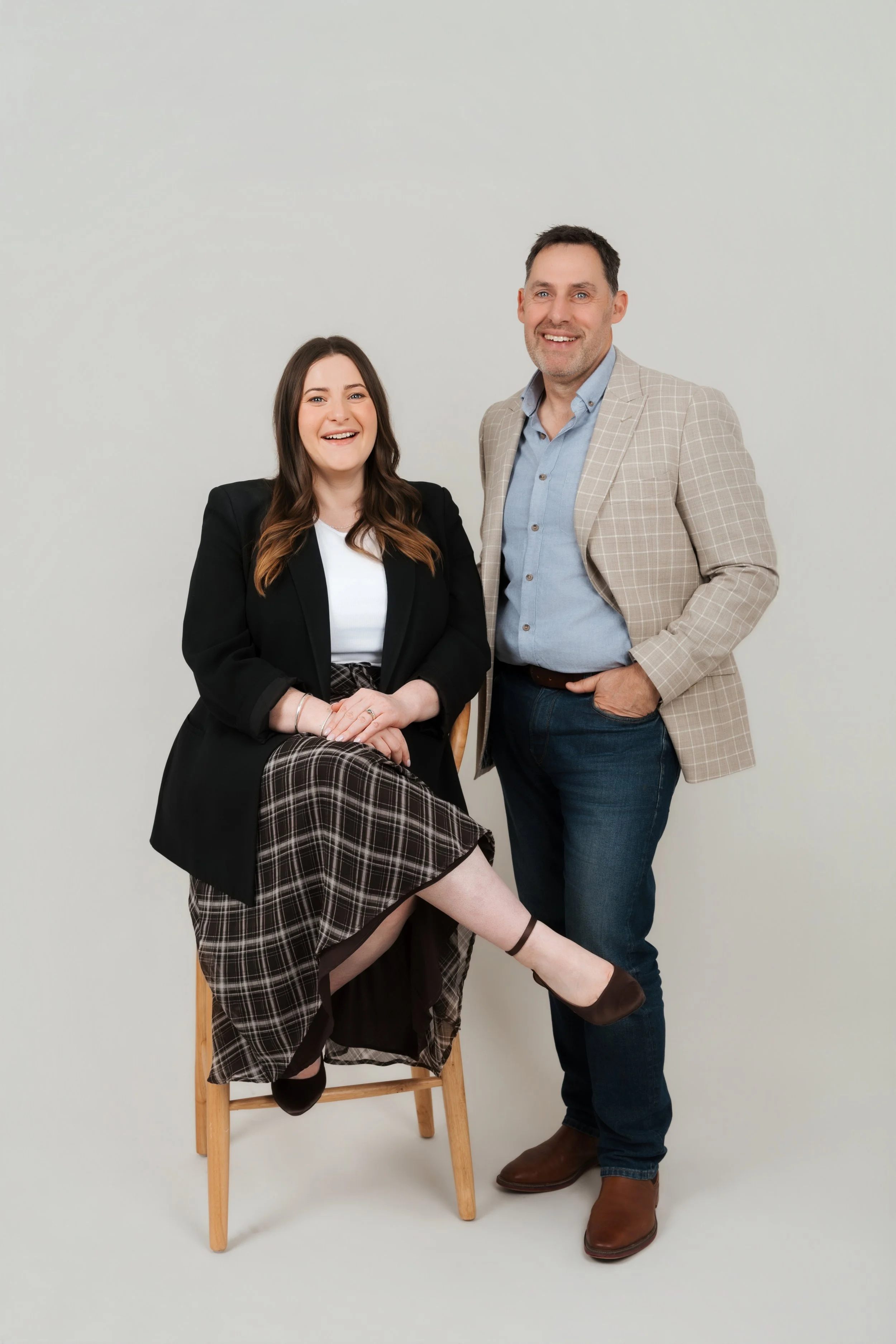 A woman with long brown hair wearing a black blazer and plaid skirt sitting on a wooden chair, smiling, with one leg crossed over the other. A man with short dark hair wearing a beige checked blazer, light blue shirt, and dark jeans stands next to her, also smiling, with one hand in his pocket. They are against a plain white background.