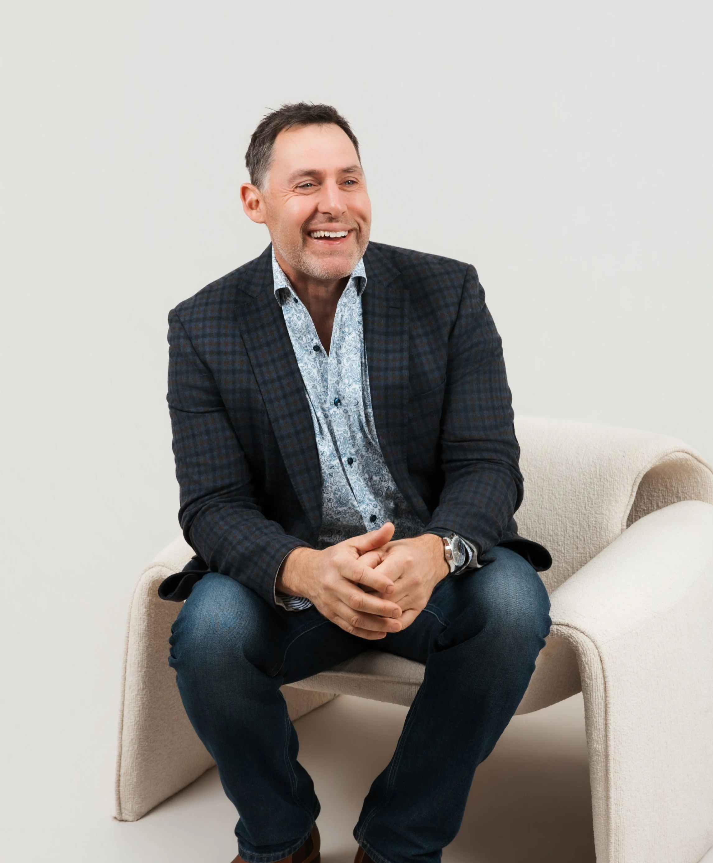 A middle-aged man with dark hair and a trimmed beard, wearing a dark plaid blazer and a light patterned shirt, sitting on a beige armchair against a plain white wall, smiling and looking to the right.