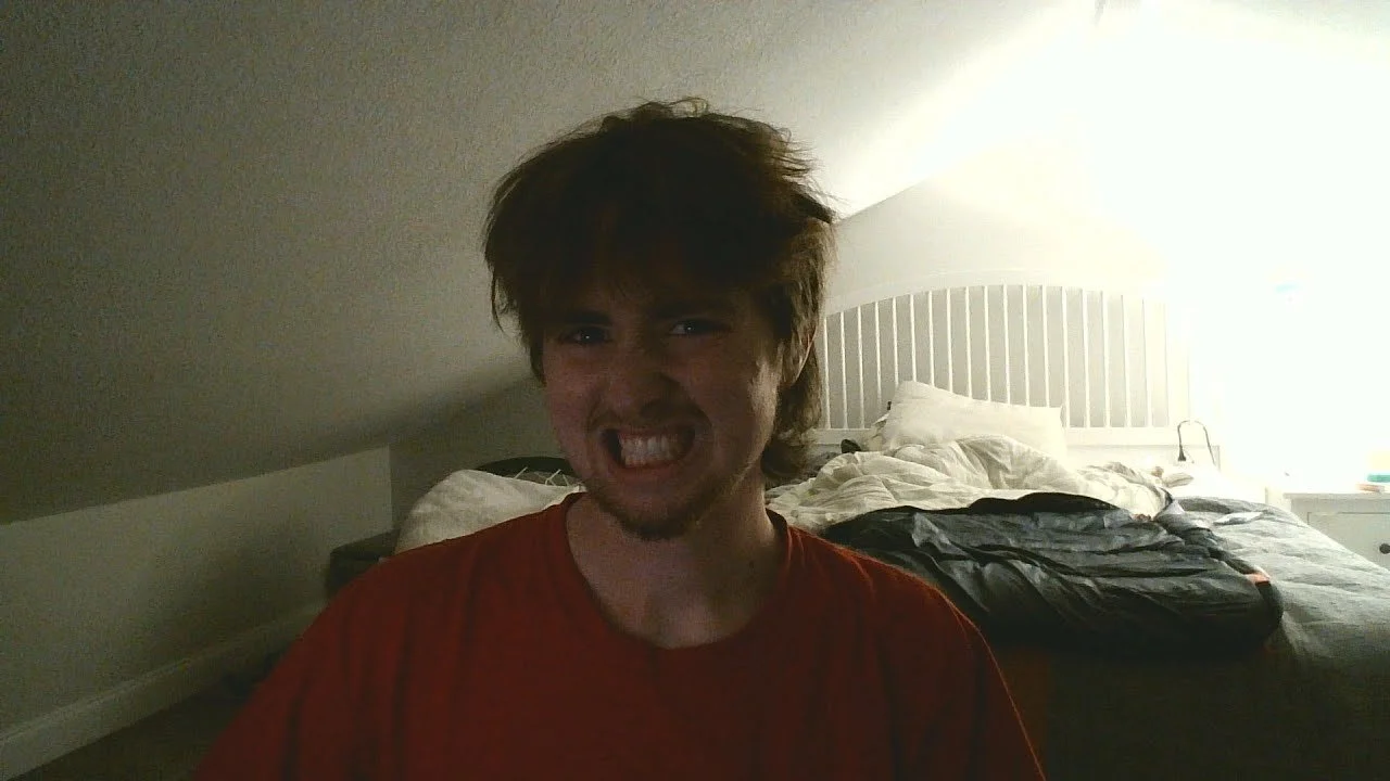 A young man with brown hair and a beard making a grimace in a bedroom with disheveled bedding and a white headboard in the background.