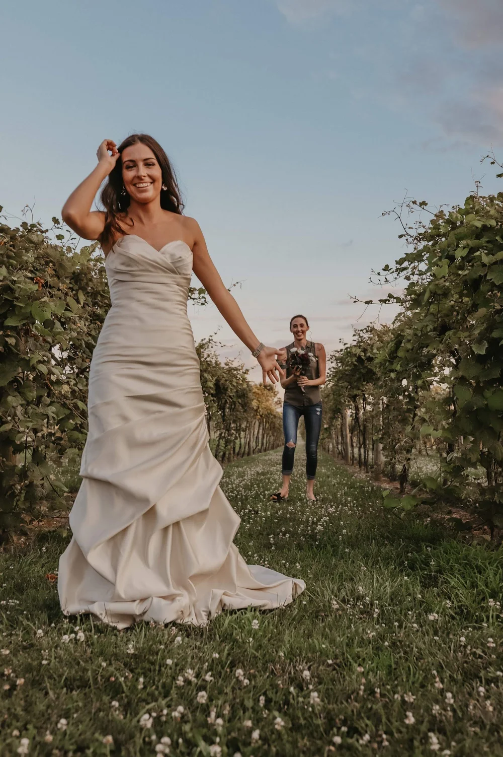 Bride in an ivory satin gown walking through a vineyard at sunset while her wedding planner holds the bouquet