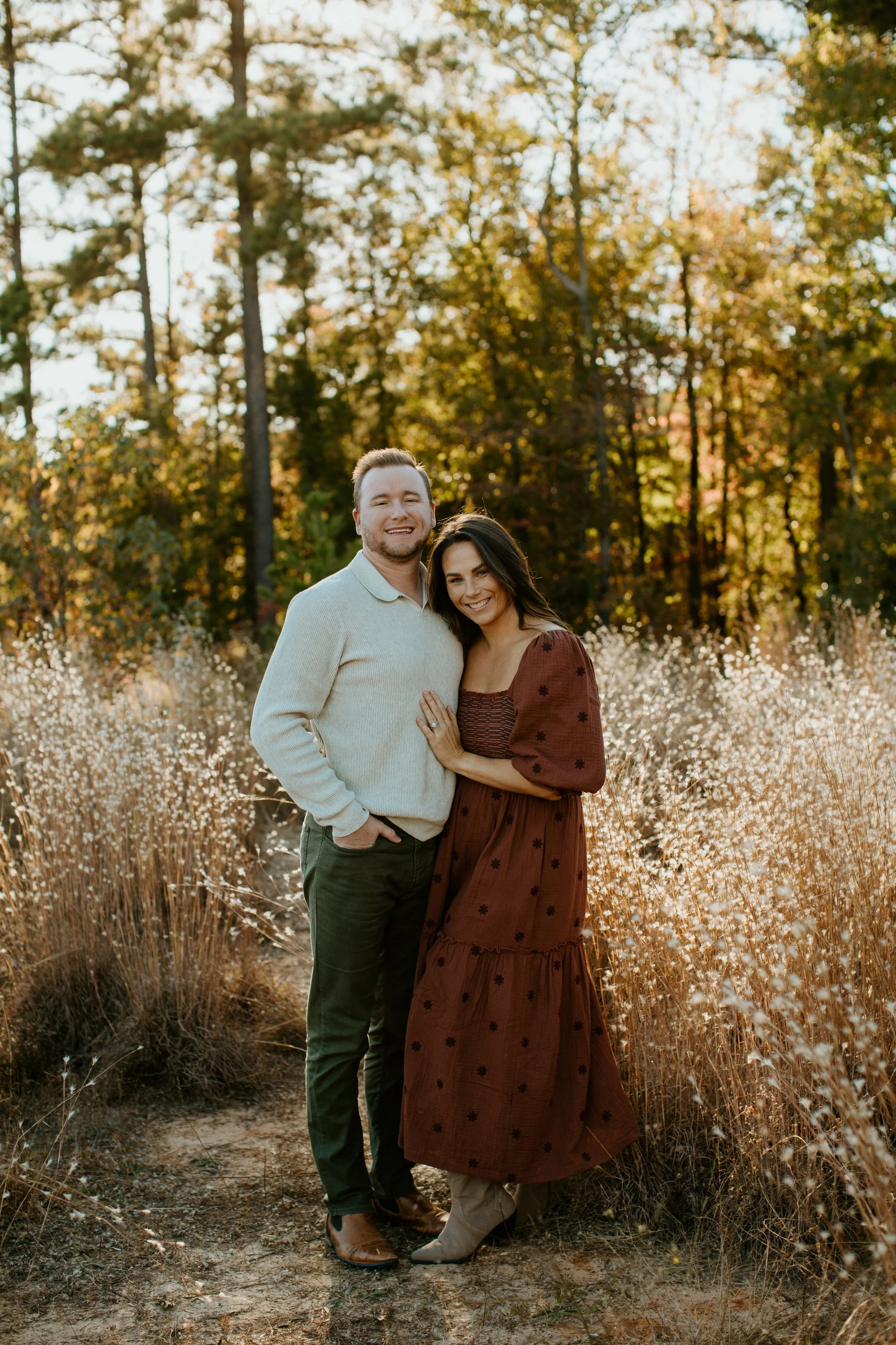 A smiling couple standing in a field of tall, dry grass with trees in the background during autumn. The man is wearing a beige sweater and green pants, and the woman is wearing a long burgundy dress with floral patterns and beige boots.
