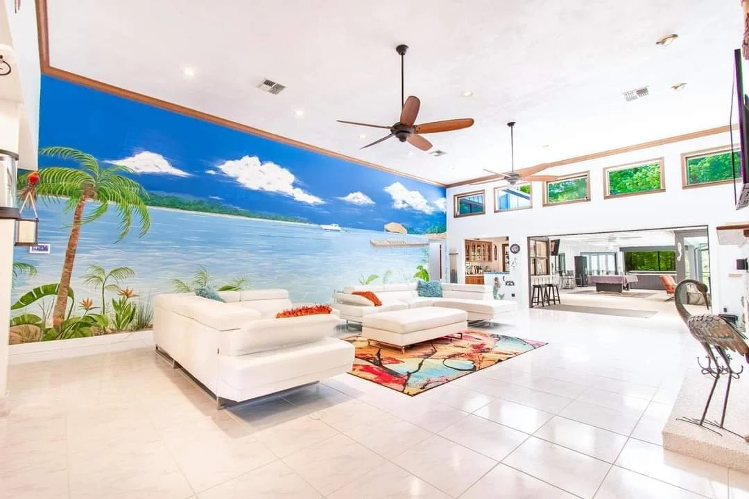 Living room with beach mural wall, white sectional sofas, ceiling fans, and tropical decor.