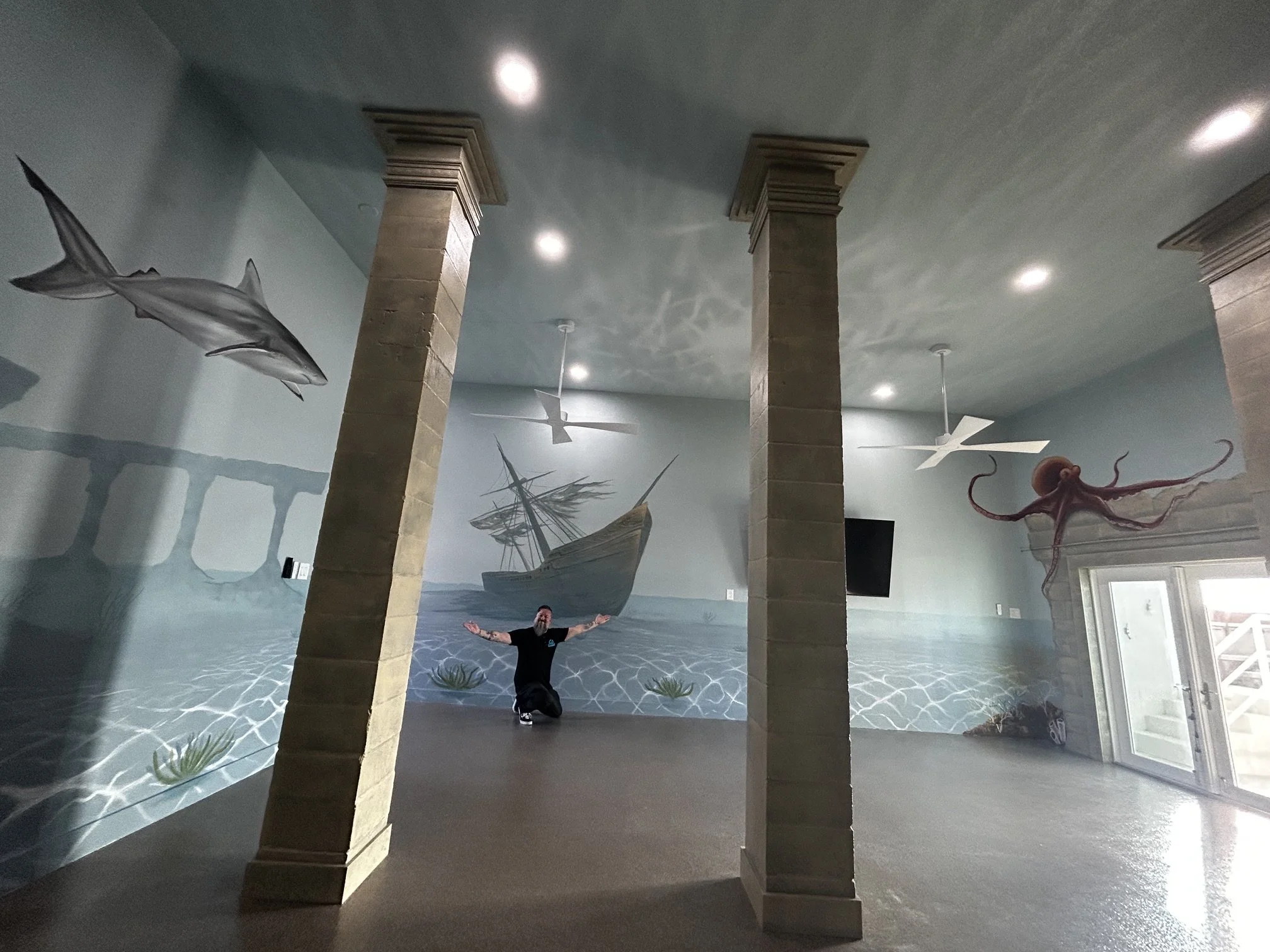 Person with arms extended, standing in front of a large, ocean-themed mural featuring a ship, sea animals, and underwater plants, with stone columns and ceiling lights.
