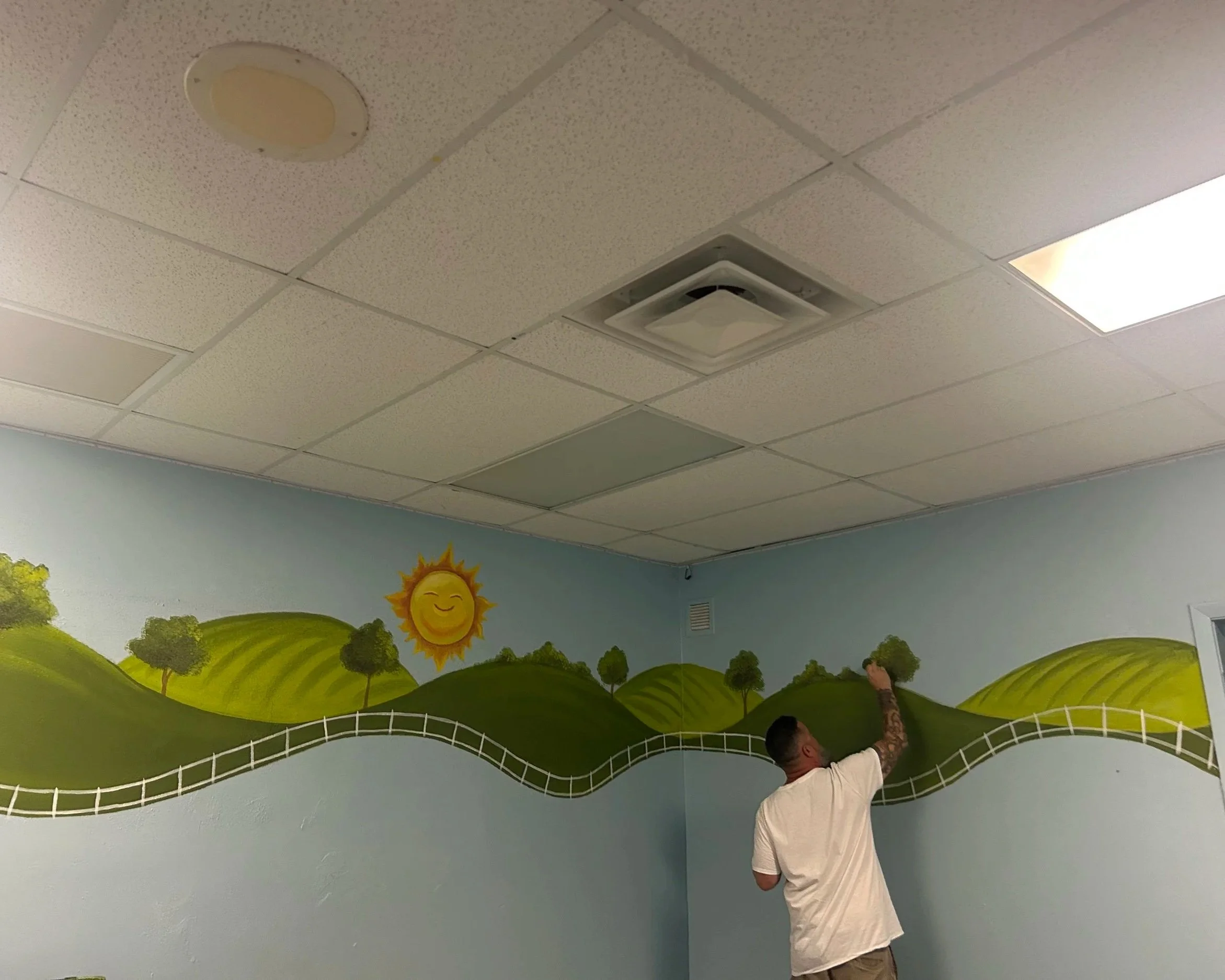 Man painting a mural of a sunny landscape with green hills, trees, and a smiling sun on an indoor wall.