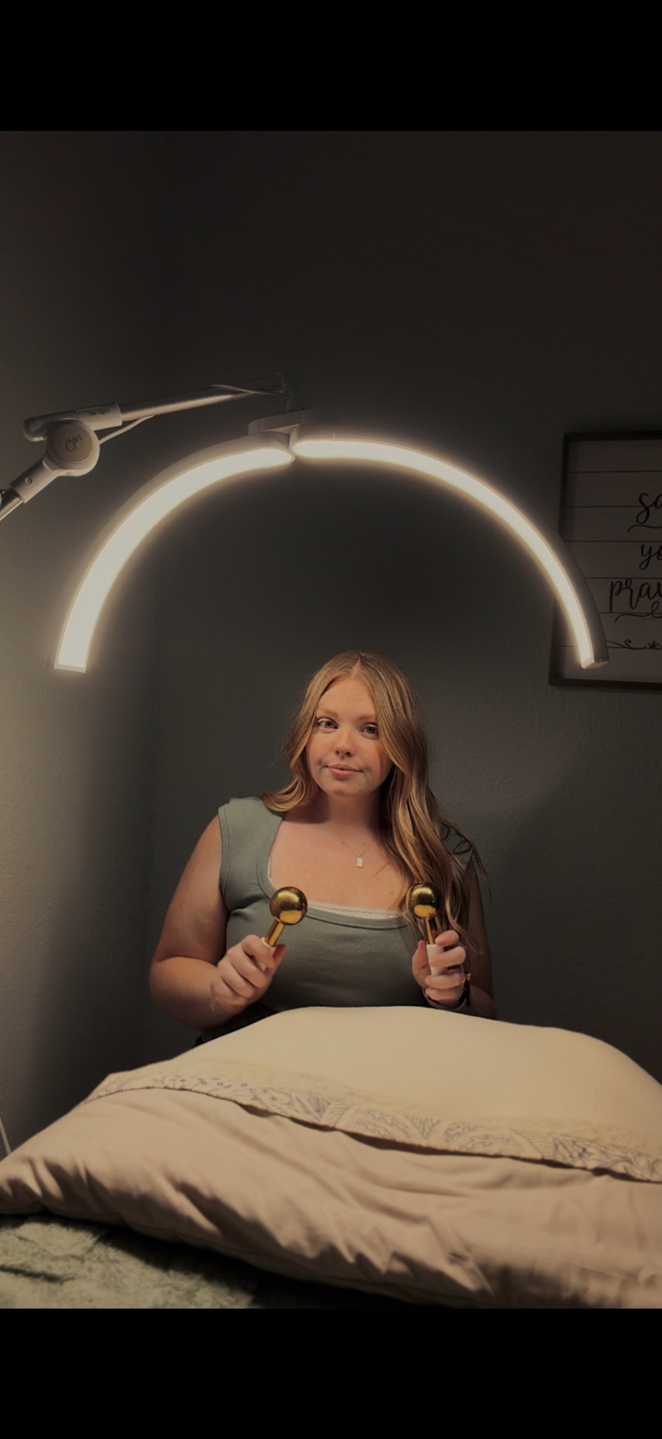 A young woman sitting in a spa or salon, holding gold massage tools, with a modern illuminated rainbow-shaped light fixture above her head.