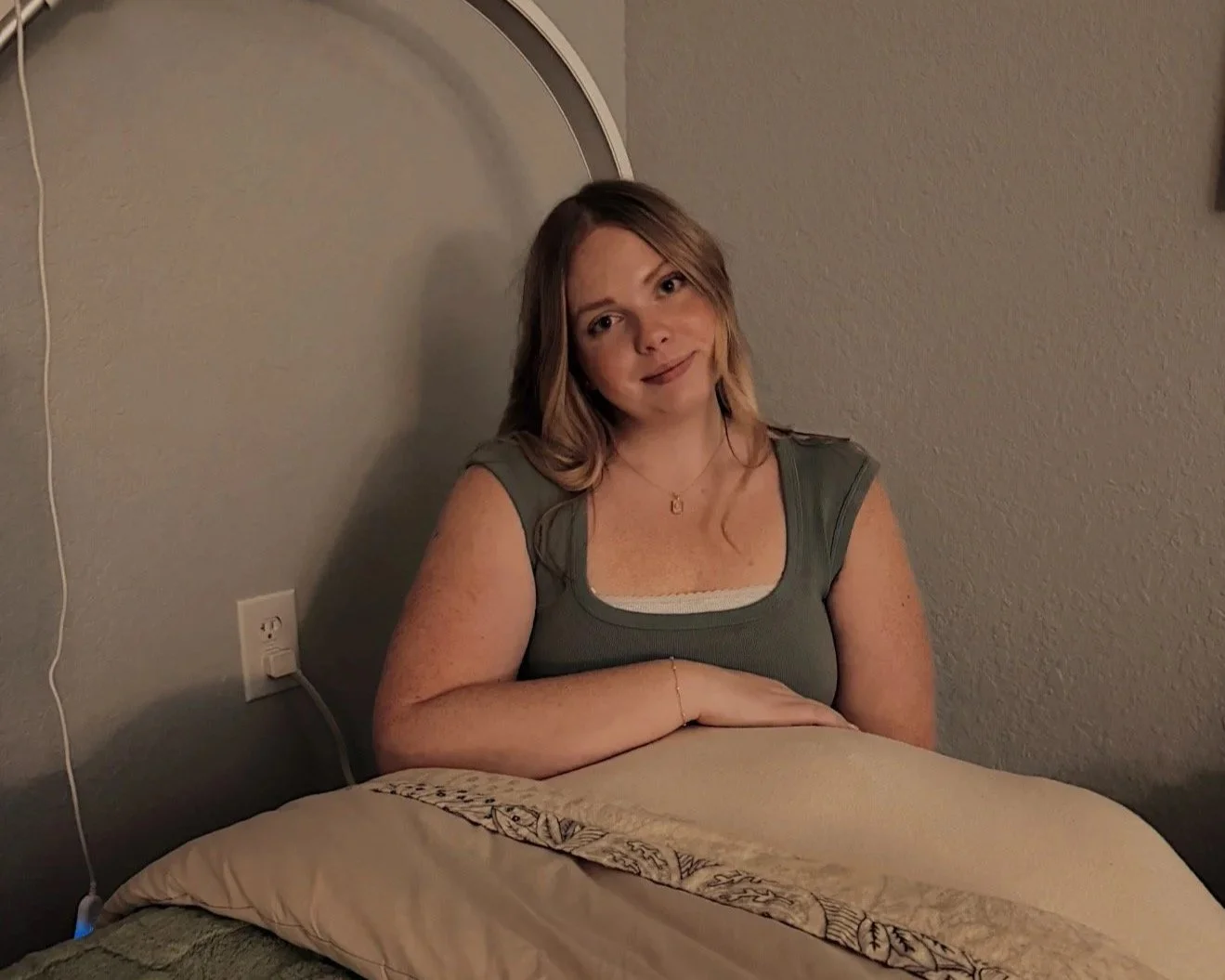 A woman with shoulder-length blonde hair smiling, sitting at a bed with her arms crossed on the bed, in a room with a plain wall and electrical outlet.