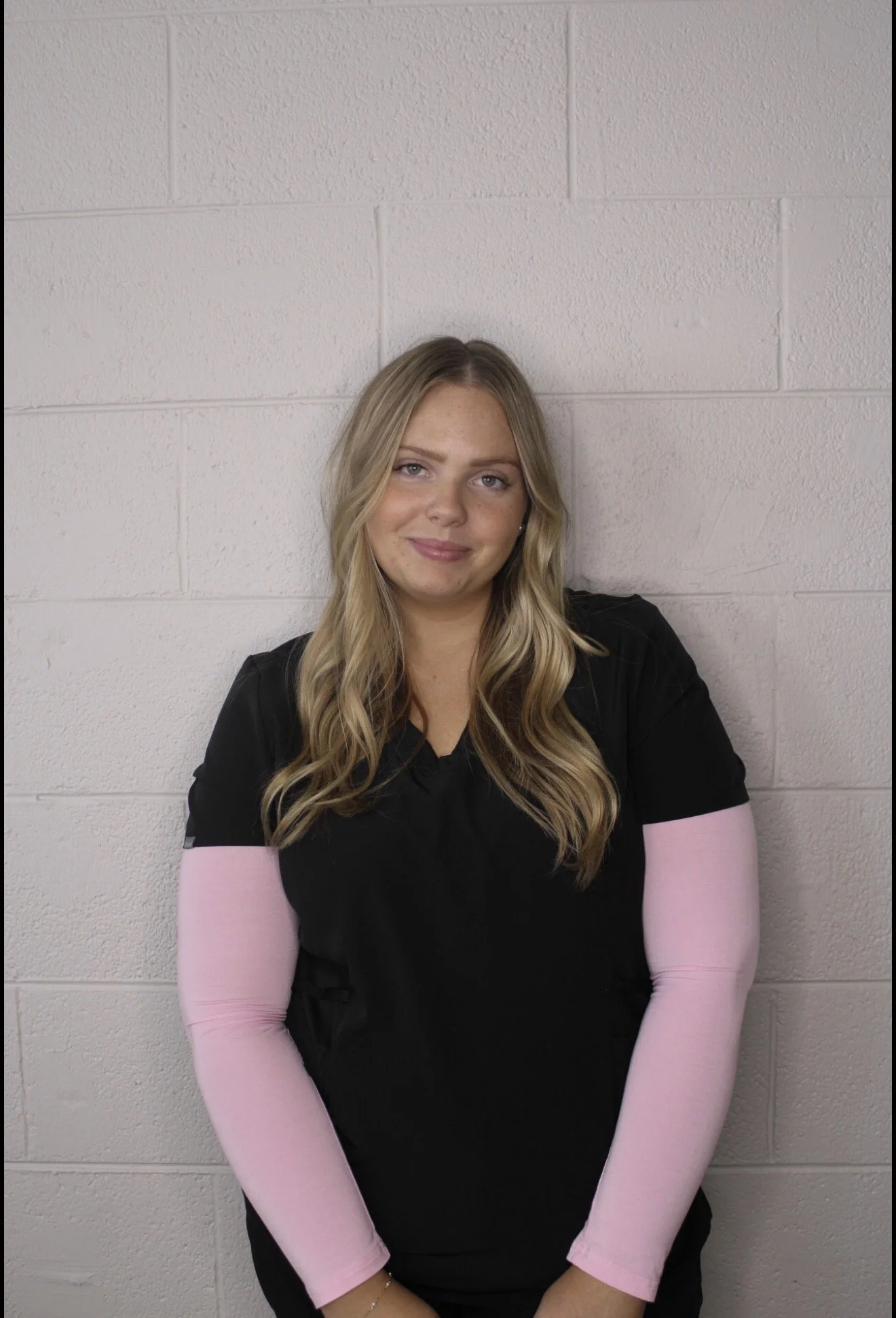 A young woman with long blonde hair wearing a black shirt and long pink arm sleeves, standing against a light-colored brick wall, smiling at the camera.