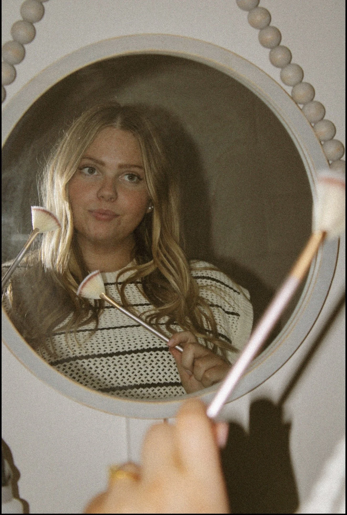 A mirror reflects a young woman with long blonde hair, holding a small brush, wearing a white sweater with black stripes.