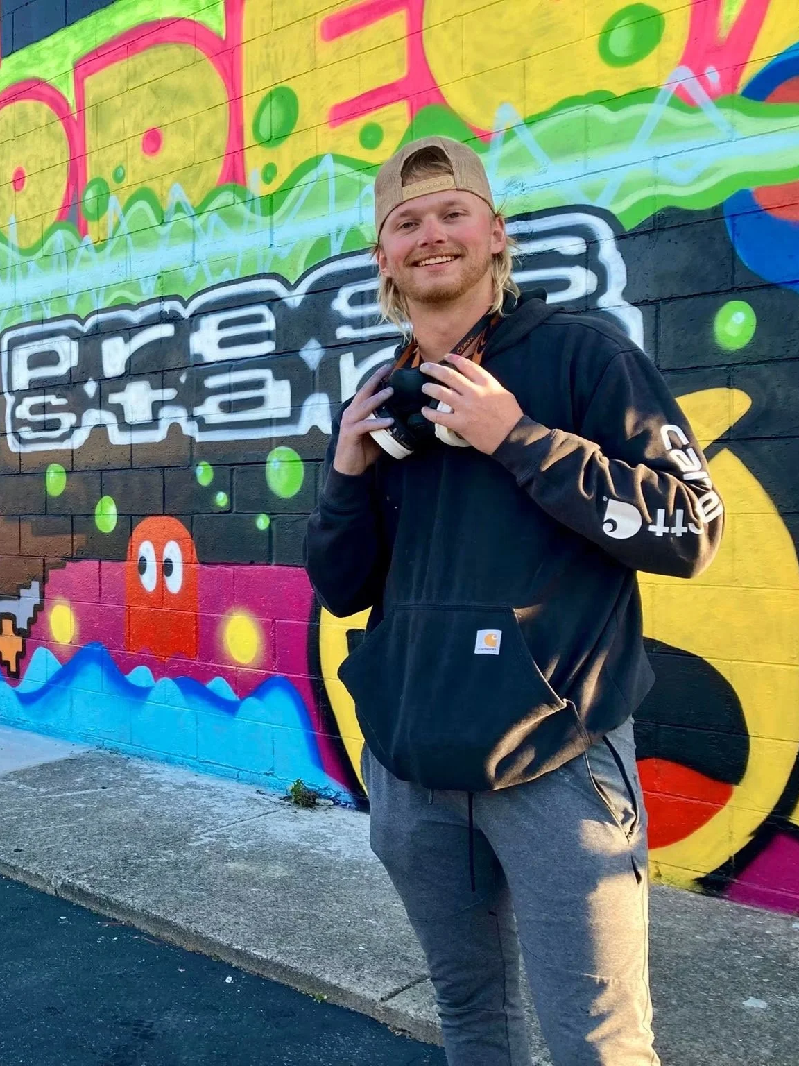 A young man with long blond hair, a beard, wearing a beige cap backwards, a black Carhartt hoodie, and gray pants, standing in front of a colorful graffiti wall smiling at the camera. He is holding a camera around his neck.