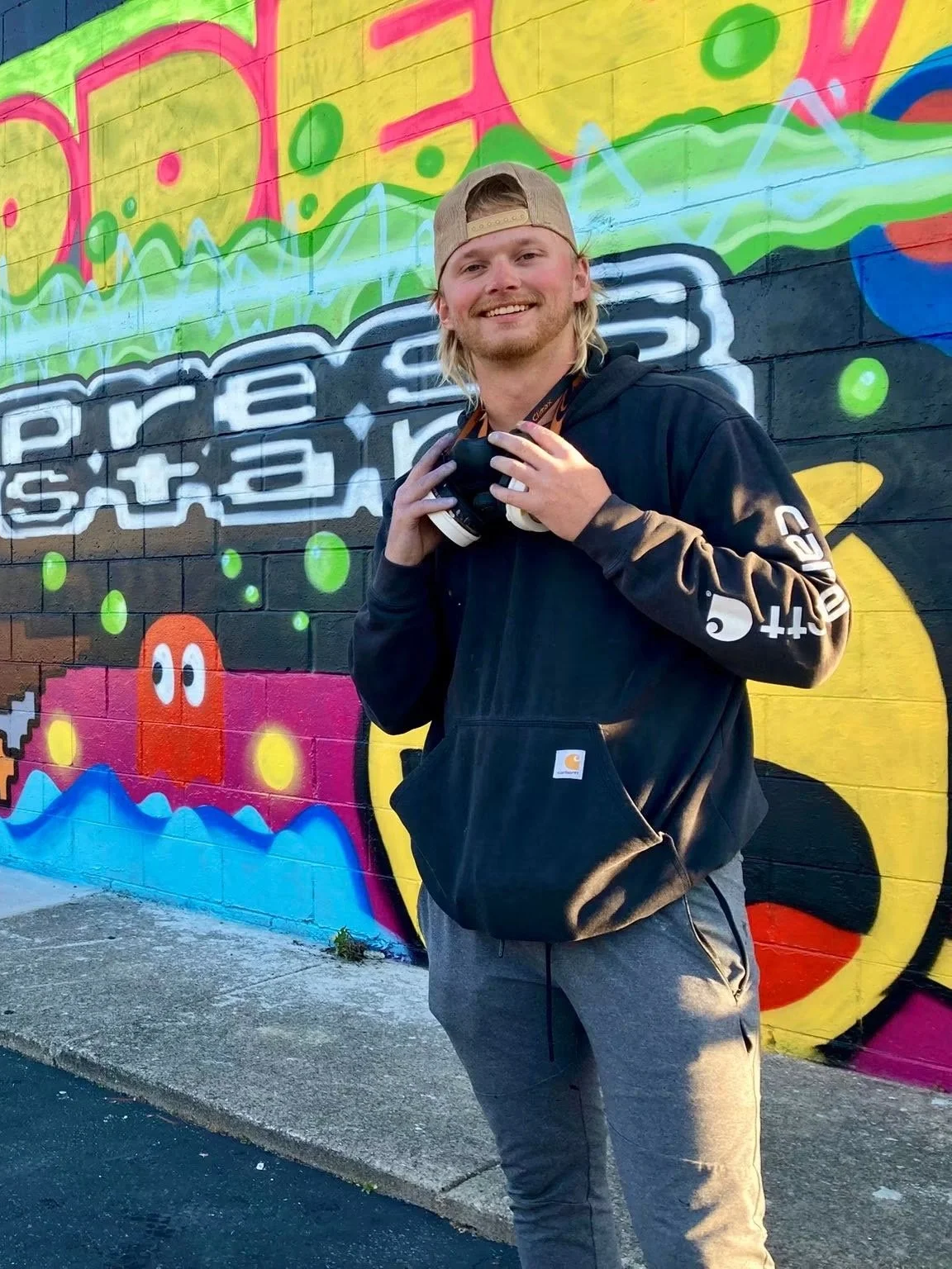 A young man standing in front of a vibrant, colorful graffiti mural on a brick wall, holding a camera around his neck and smiling at the camera.