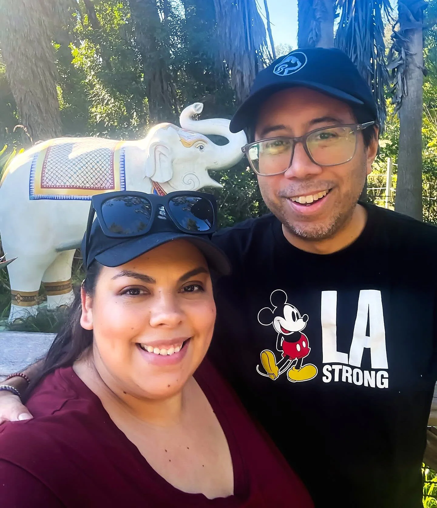 A smiling man and woman taking a selfie outdoors with a decorated white elephant statue in the background.