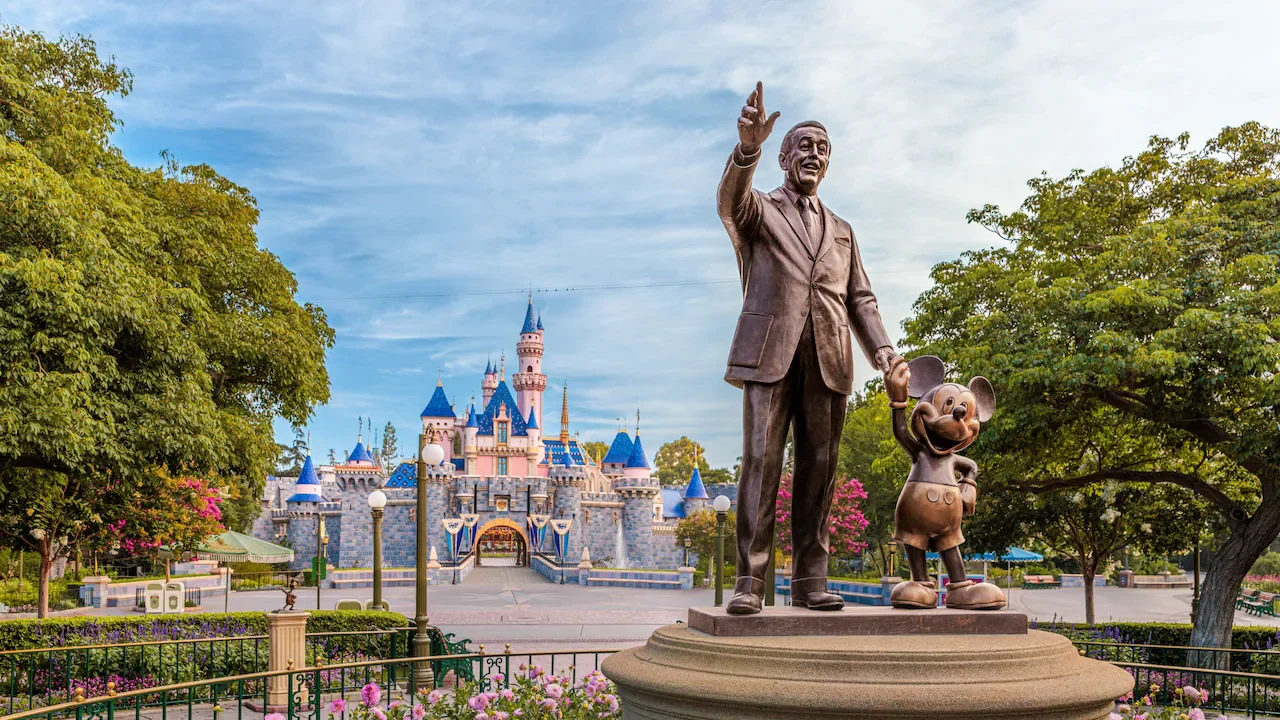 Statue of Walt Disney holding Mickey Mouse's hand in front of Disneyland castle with trees and pink flowers.