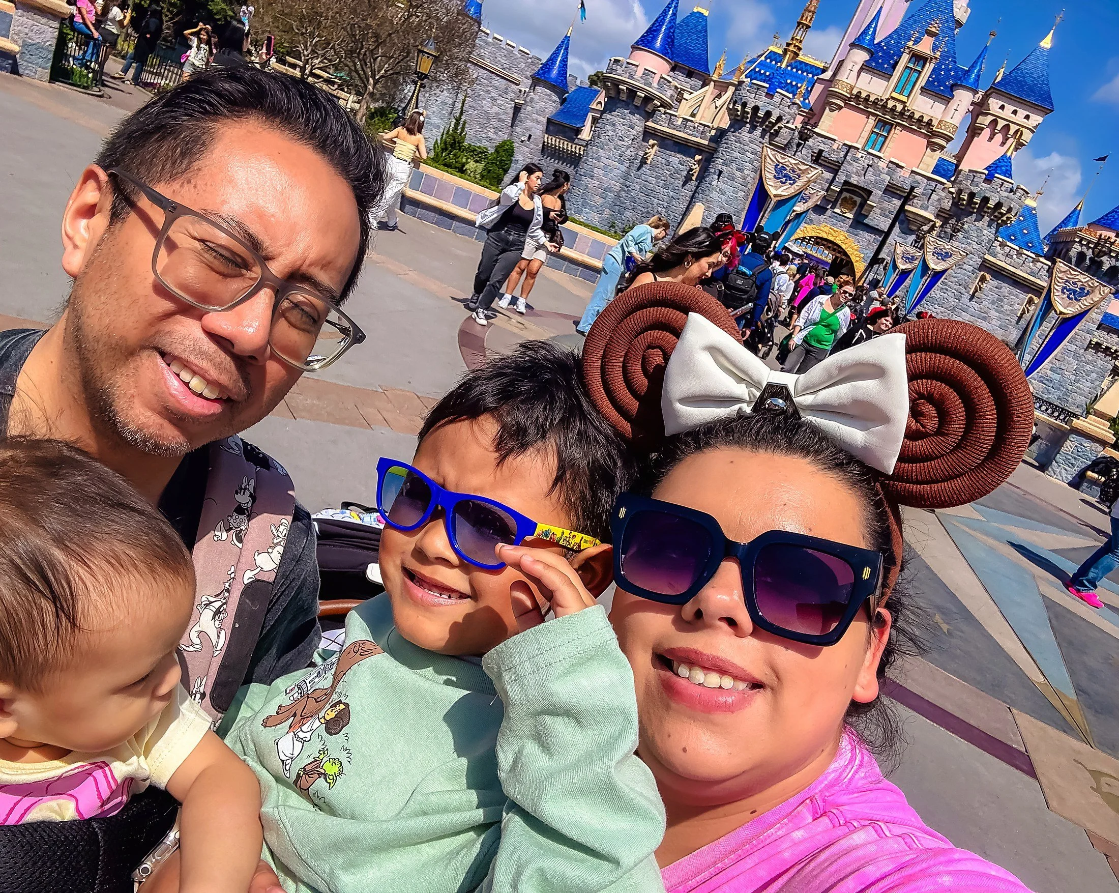 A group of people, including a man with glasses, several children, and a woman, taking a selfie in front of Disneyland's Sleeping Beauty Castle on a sunny day.