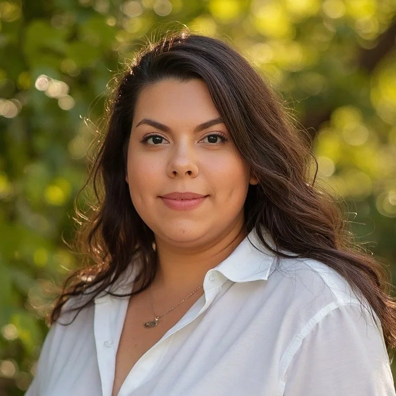 A woman with long dark hair wearing a white button-up shirt outdoors, with a blurred green background and sunlight.