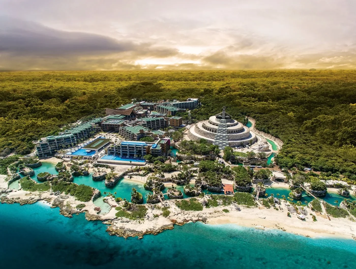 An aerial view of a large tropical resort with multiple buildings, swimming pools, and a pagoda-like structure surrounded by lush green forest and coastline.