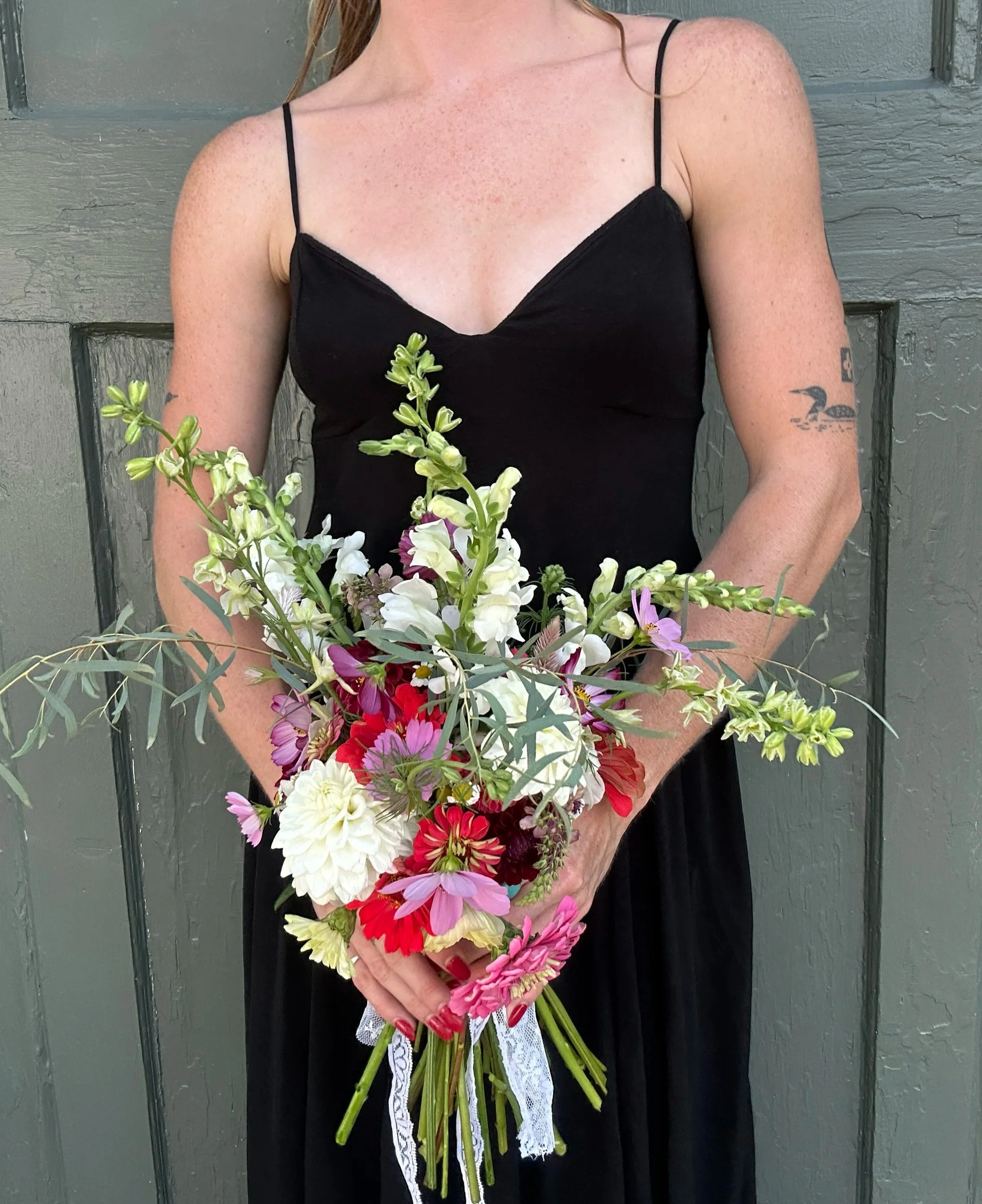 A woman in a black dress holding a bouquet of mixed flowers, standing in front of a gray wooden door. The woman has freckles, a tattoo on her left arm, and red hair.