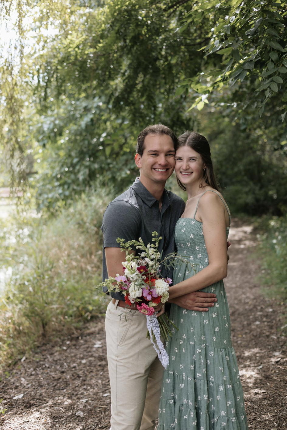 A young couple standing on a dirt path in a wooded area, smiling, with the woman holding a colorful bouquet of flowers.