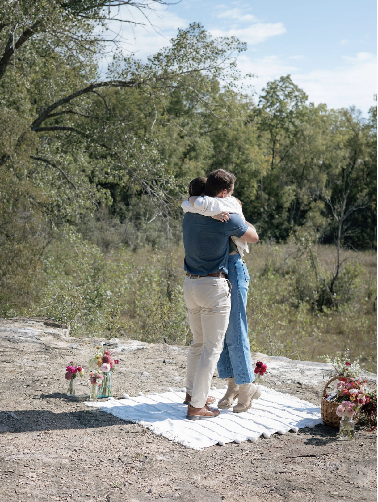 A couple hugging during a wedding ceremony outdoors on a rocky area with trees in the background. A white cloth and flower vases are on the ground nearby.